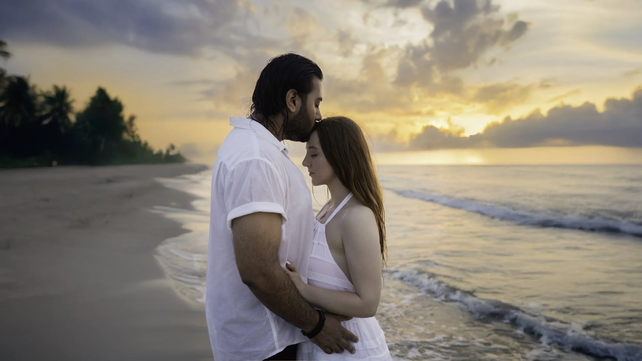 A couple standing close together on a beach at sunset, with the man gently resting his forehead on the woman's, both with their eyes closed, the beach and ocean in the background.