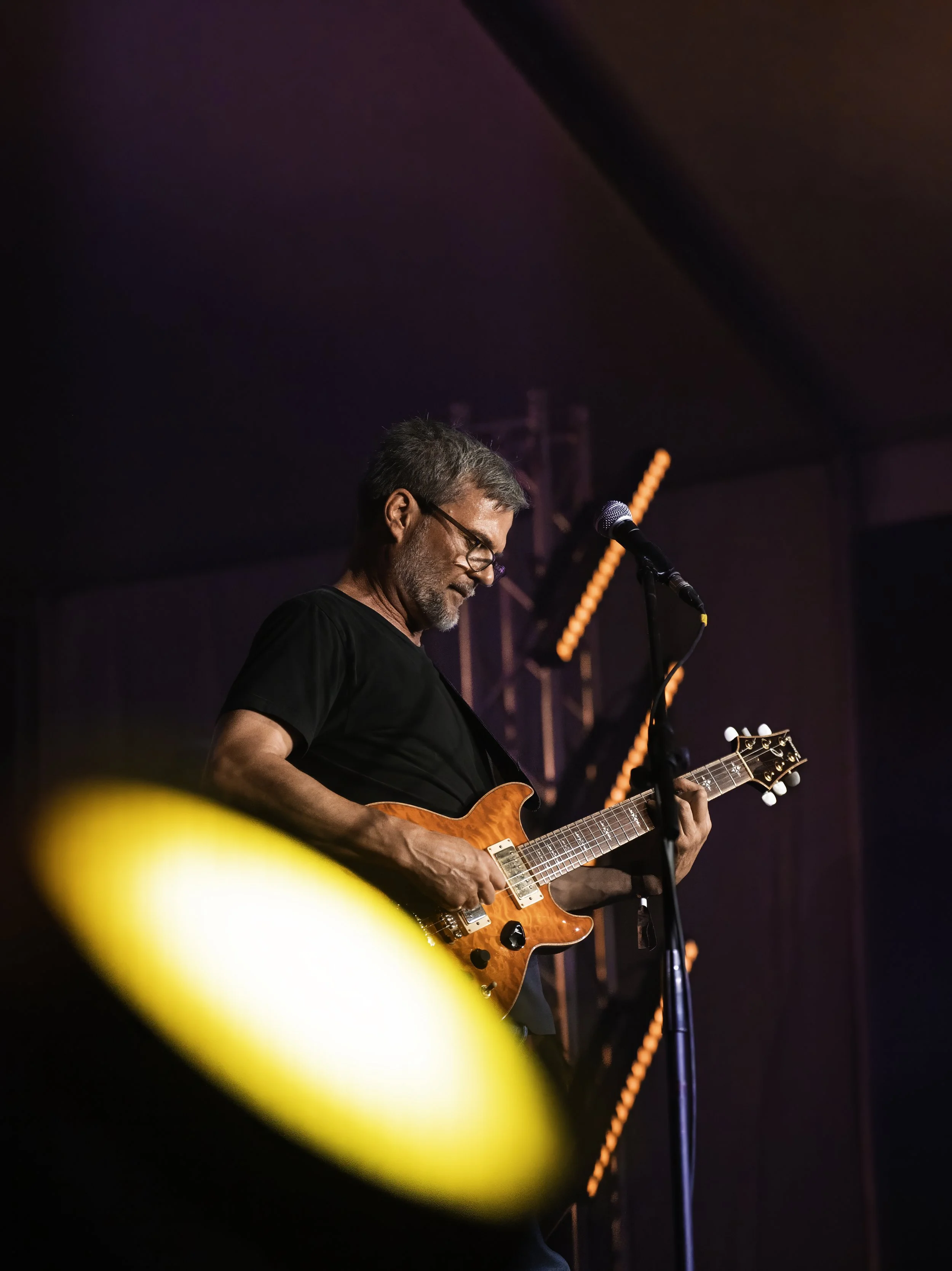 A man with gray hair and glasses playing an orange electric guitar on stage. The stage has purple and orange lighting effects with visible equipment and a microphone stand.