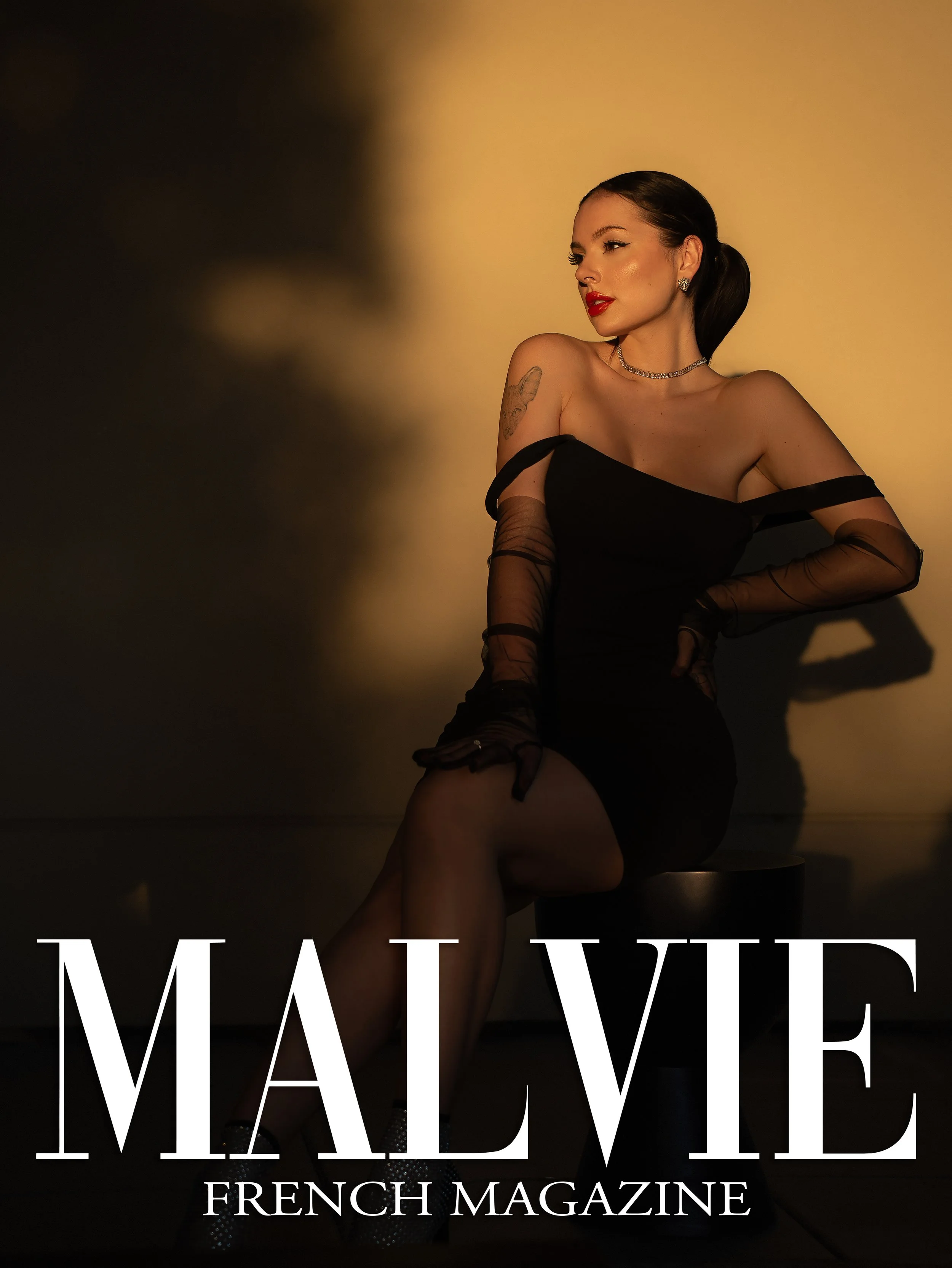 Woman with dark hair in an elegant off-shoulder black dress, standing in front of a beige background, with makeup and jewelry; overlaid with the text 'MAY VIBE' and 'FRENCH MAGAZINE' at the bottom.