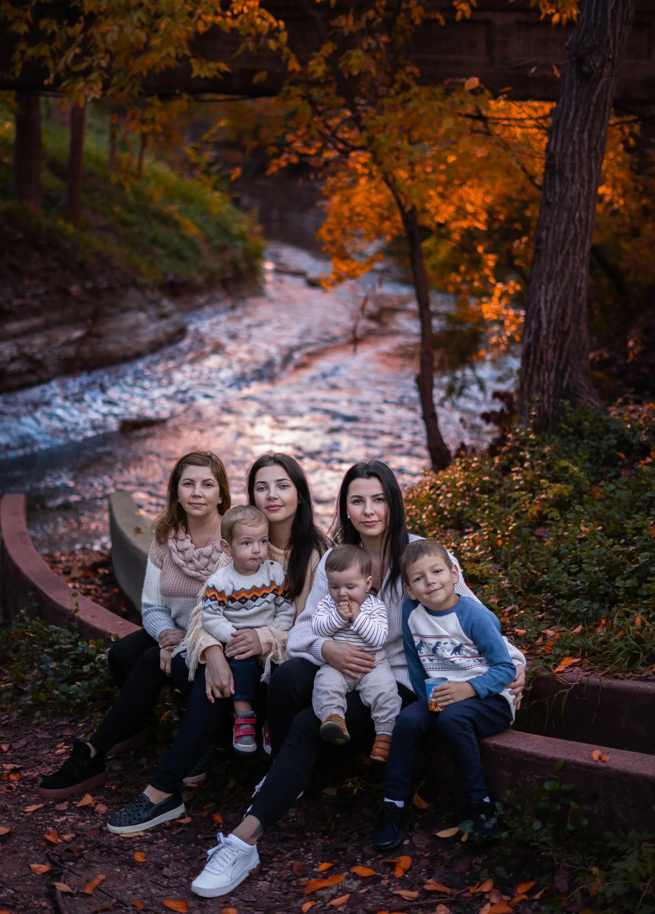 A family group of seven people, including adults and children, sitting on a curved park bench outdoors during autumn. In the background, there are trees with fall foliage and a stream flowing underneath a bridge.