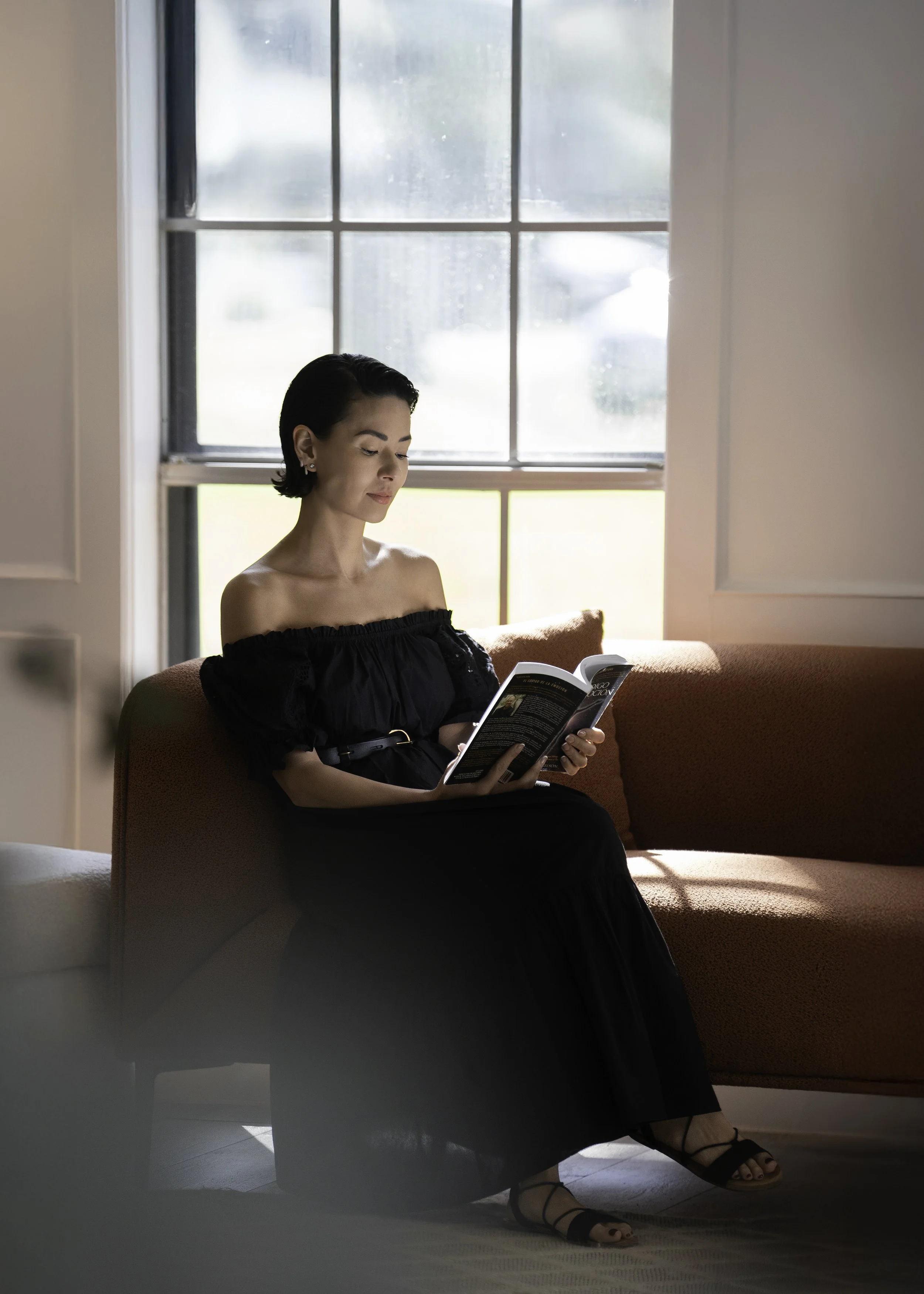 A woman with short dark hair, wearing a black off-shoulder dress and black sandals, sitting on a pinkish-orange sofa reading a magazine. She is near a large window with divided panes, allowing natural light to illuminate her and the room.