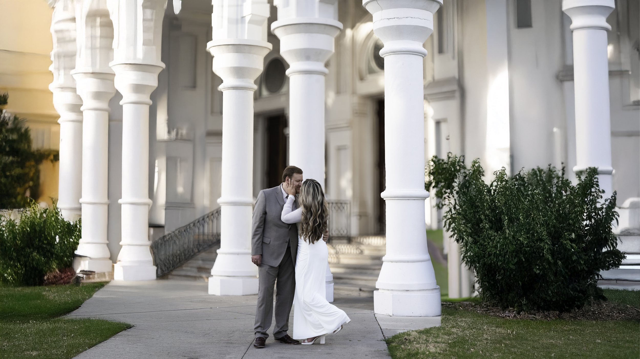 A couple dressed in wedding attire sharing an intimate moment outside a white building with large columns and greenery.