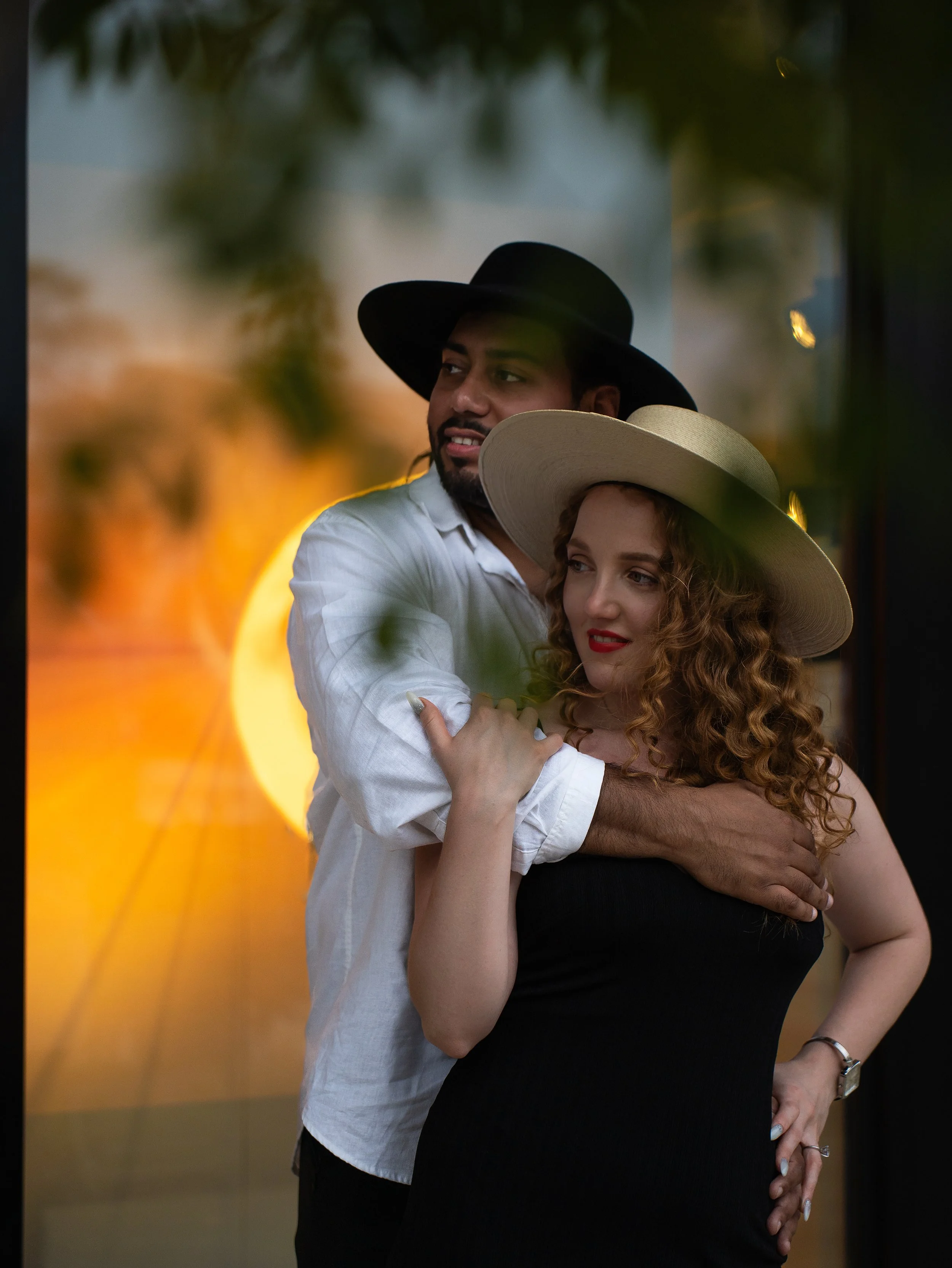 A man and woman embracing, both wearing wide-brimmed hats, with a blurred sunset in the background.