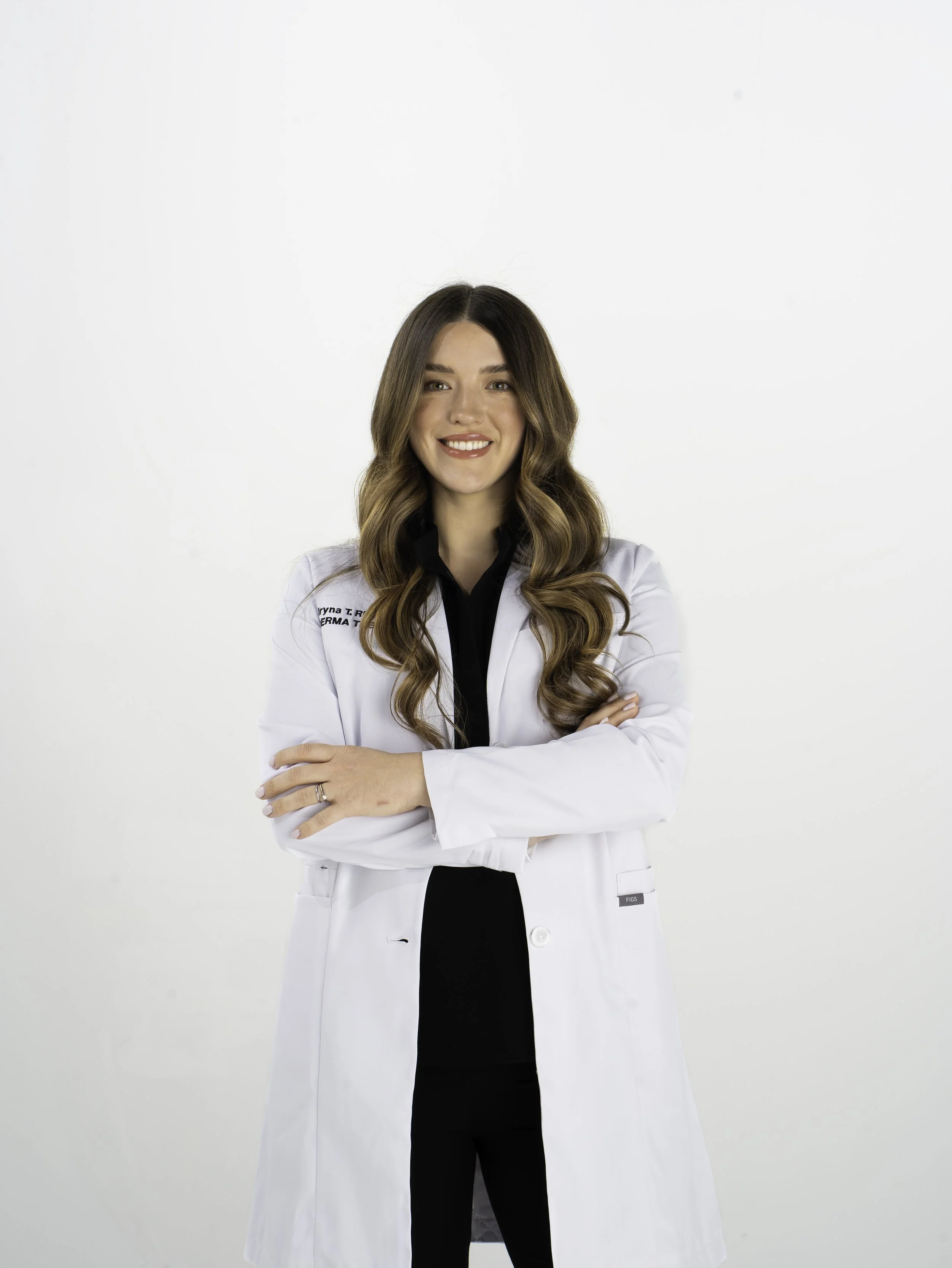 A woman with wavy brown hair smiling and standing with arms crossed. She is wearing a white medical coat over black clothing, against a plain white background.