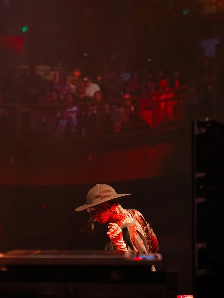 A DJ wearing a wide-brimmed hat, bandana, and gloves performing at a nightclub with a crowd on an upper balcony watching.