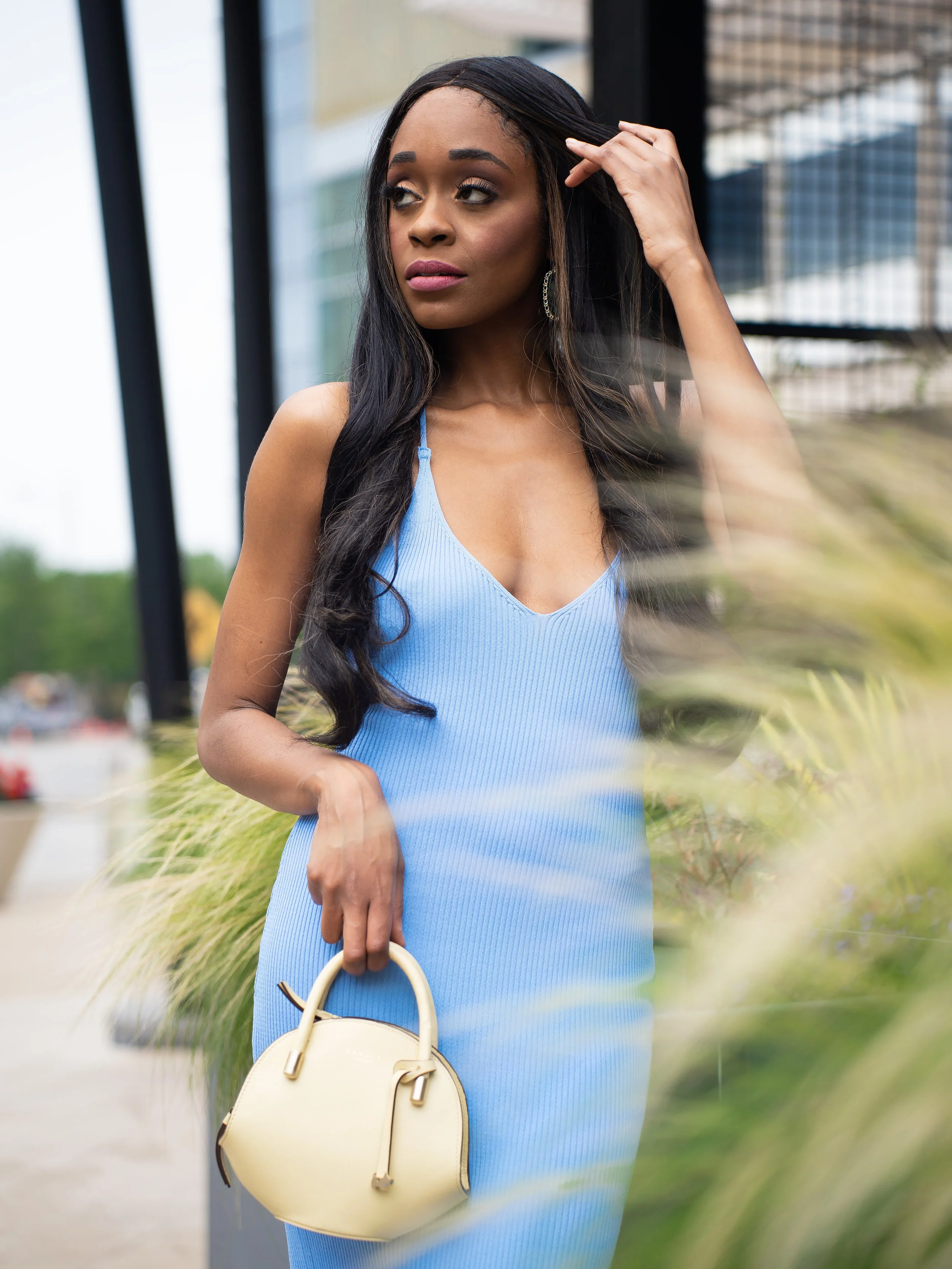 A woman in a blue dress holding a beige handbag, standing outdoors near modern glass buildings, with her hand touching her hair and looking to the side.