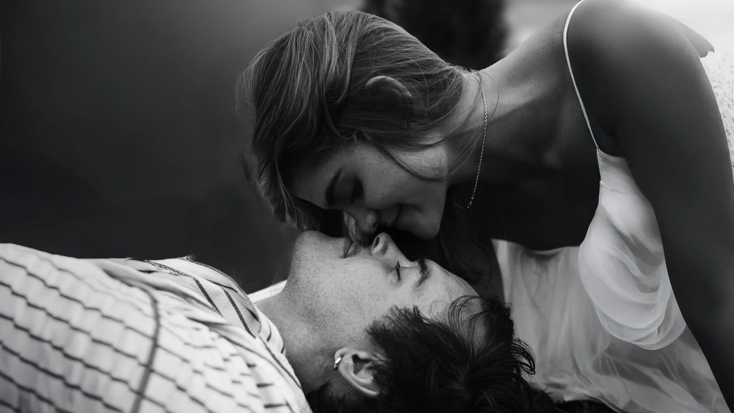 A woman leans over a man lying in bed, seemingly about to kiss him, in black and white.
