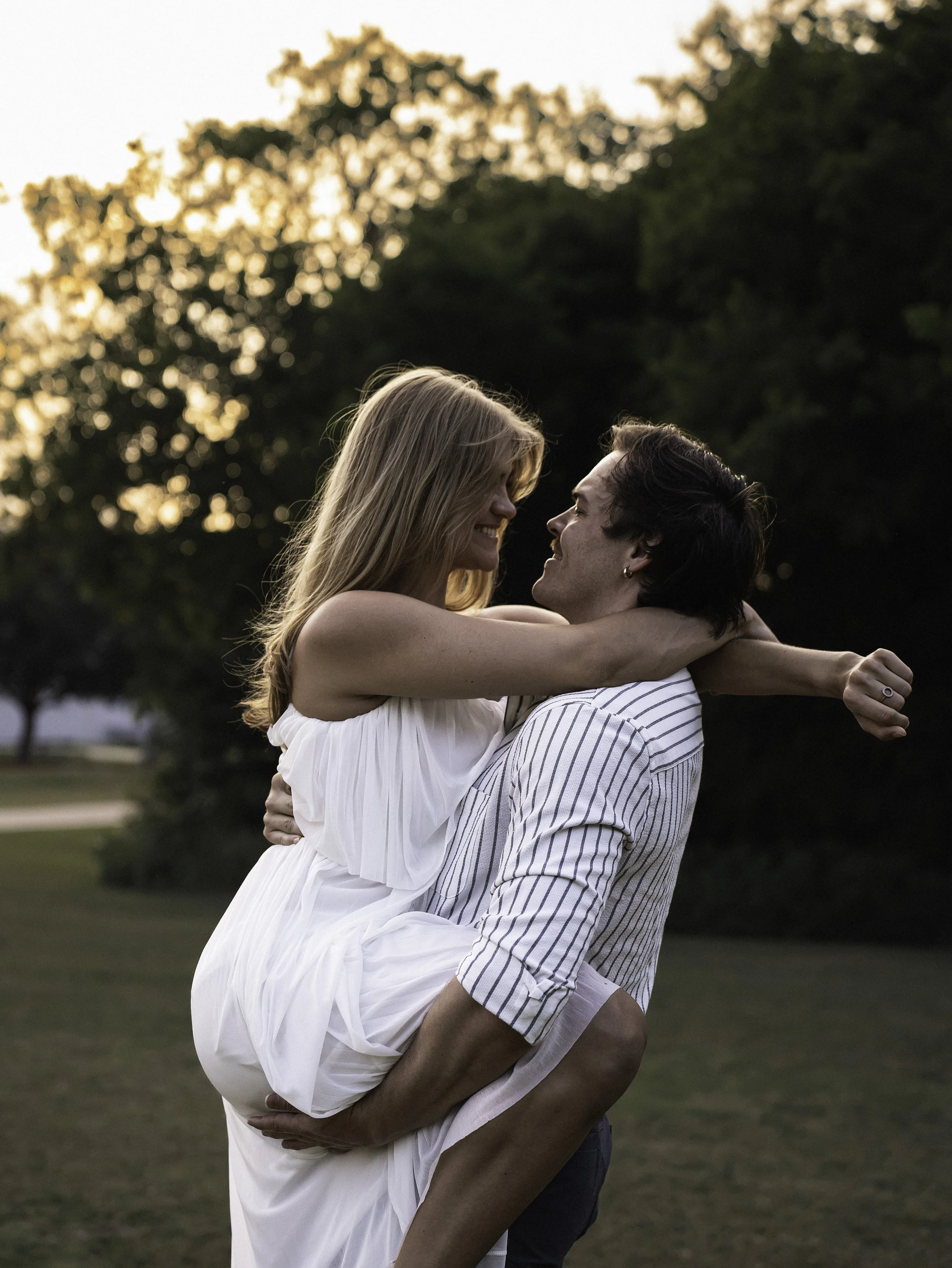 A couple is dancing outdoors during sunset, with trees and a grassy area in the background.