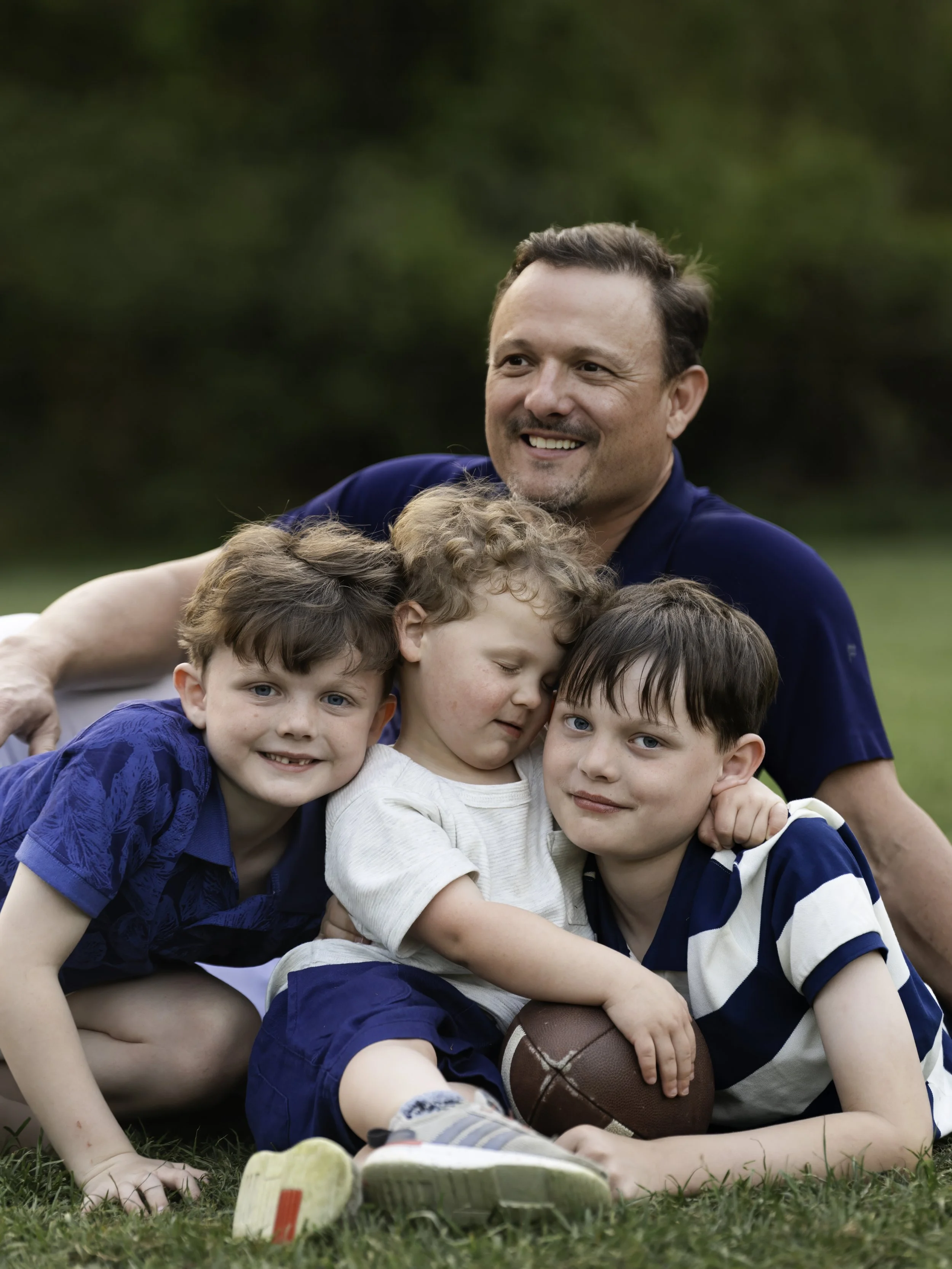 A man with short brown hair smiling and hugging a group of children on the grass. The kids are sitting or lying down, holding a football, and looking at the camera. The background shows blurred green foliage.