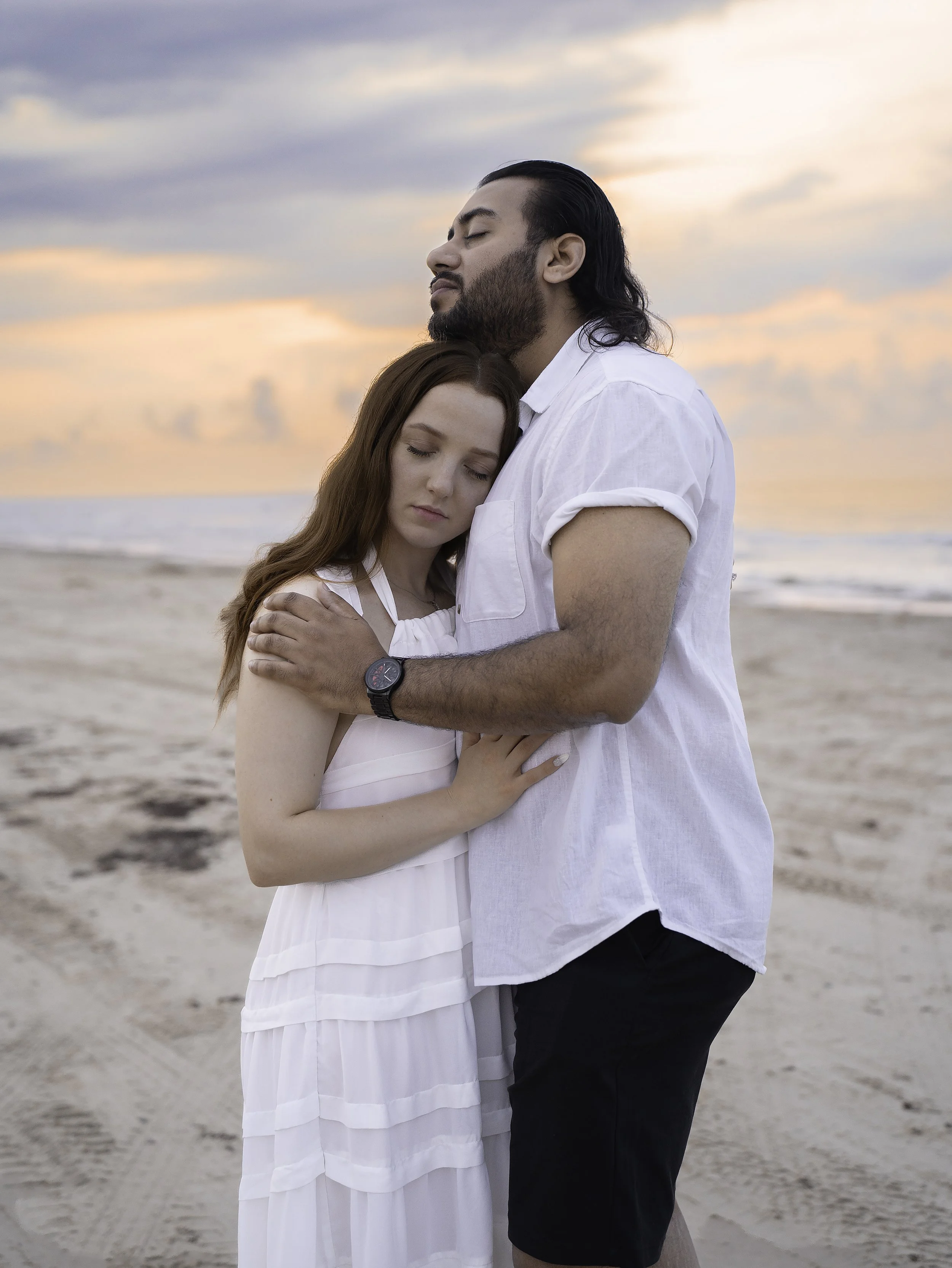 A man and woman embrace on a beach at sunset, with the man gently resting his head on the woman's head and both with eyes closed.
