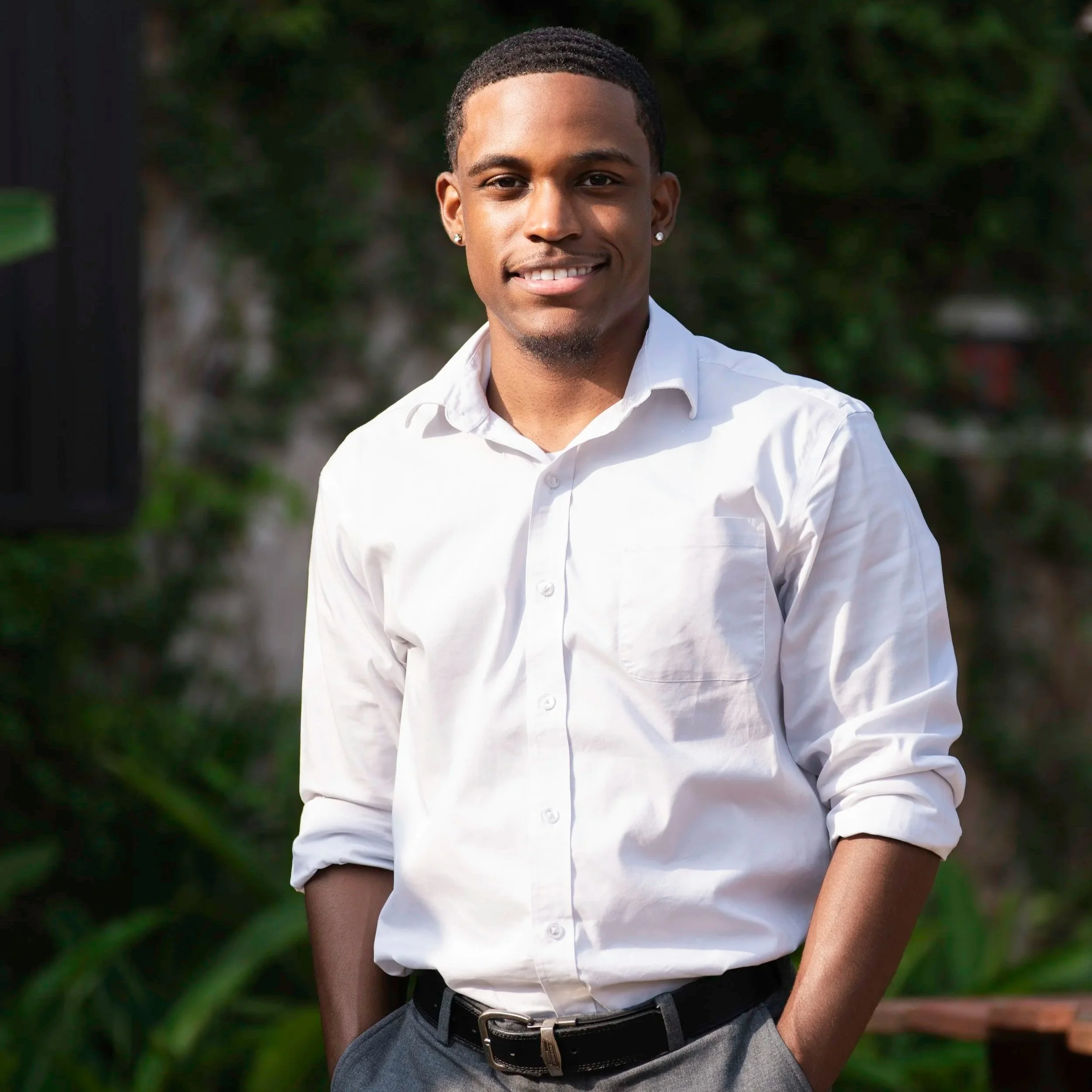 A young man with a short haircut and earrings, dressed in a white button-up shirt with rolled-up sleeves, standing outdoors with a blurred greenery background, smiling at the camera.