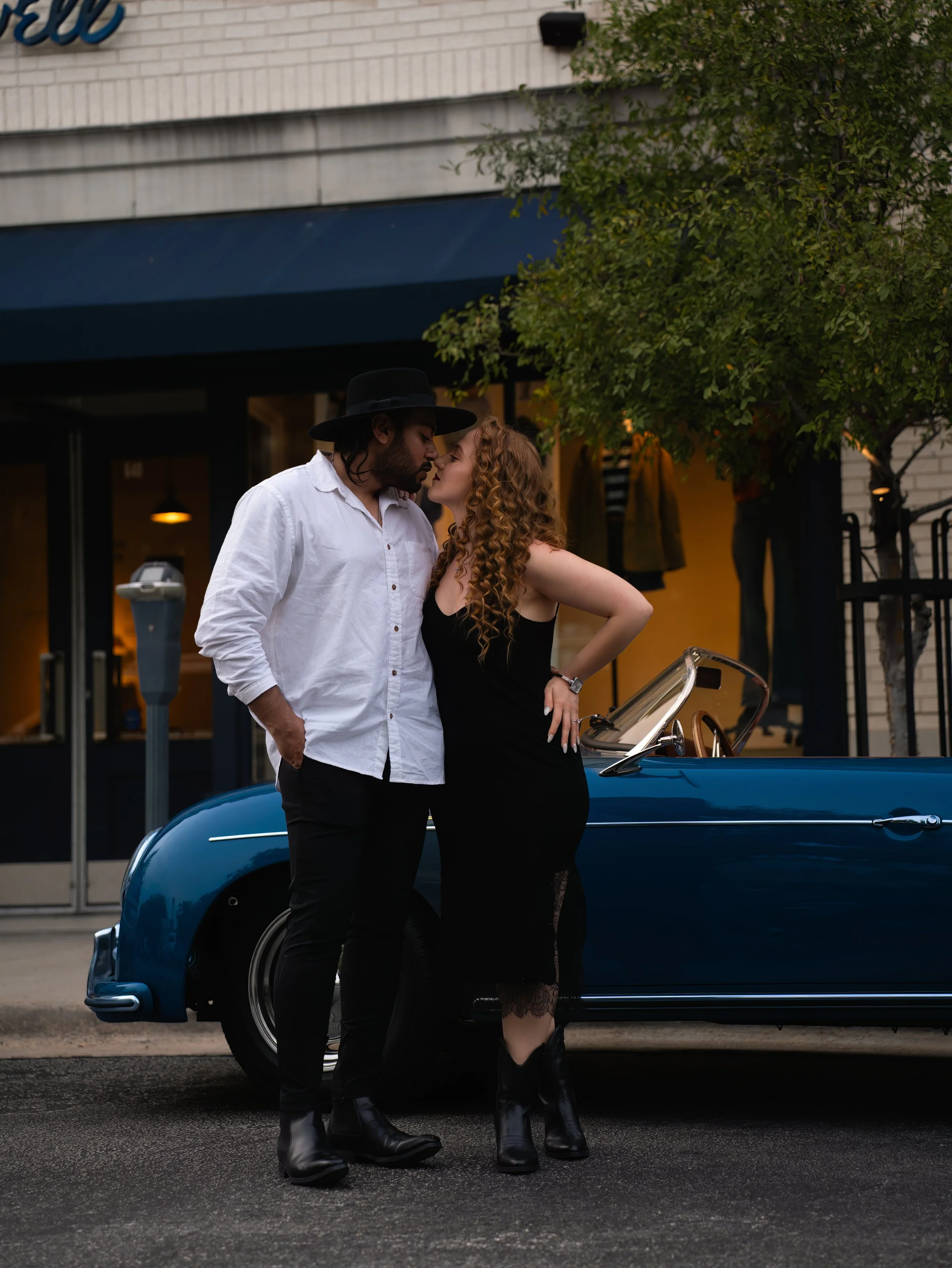 A couple stands closely in front of a vintage blue convertible car, about to kiss, on a city street during twilight. The man wears a white shirt, black fedora, and black boots; the woman wears a black dress and lace stockings.