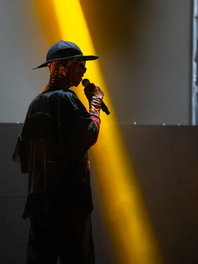 Person wearing a hat and sunglasses, holding a microphone, standing in front of a brightly lit background.