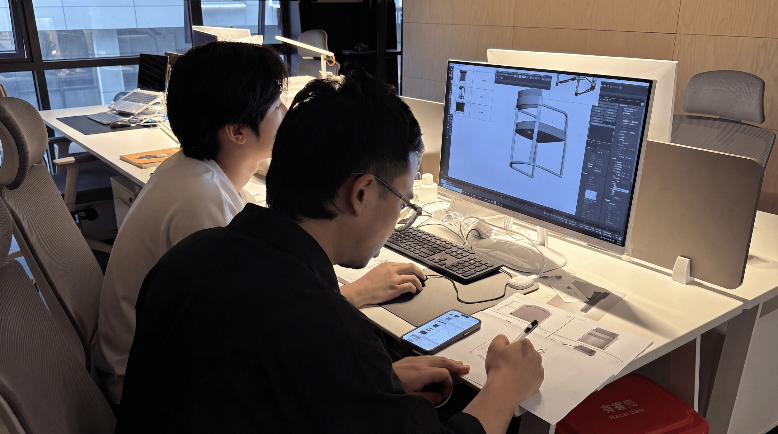 Two people working on a design project at a desk in an office, with a large computer monitor displaying a 3D design of a chair or furniture piece.