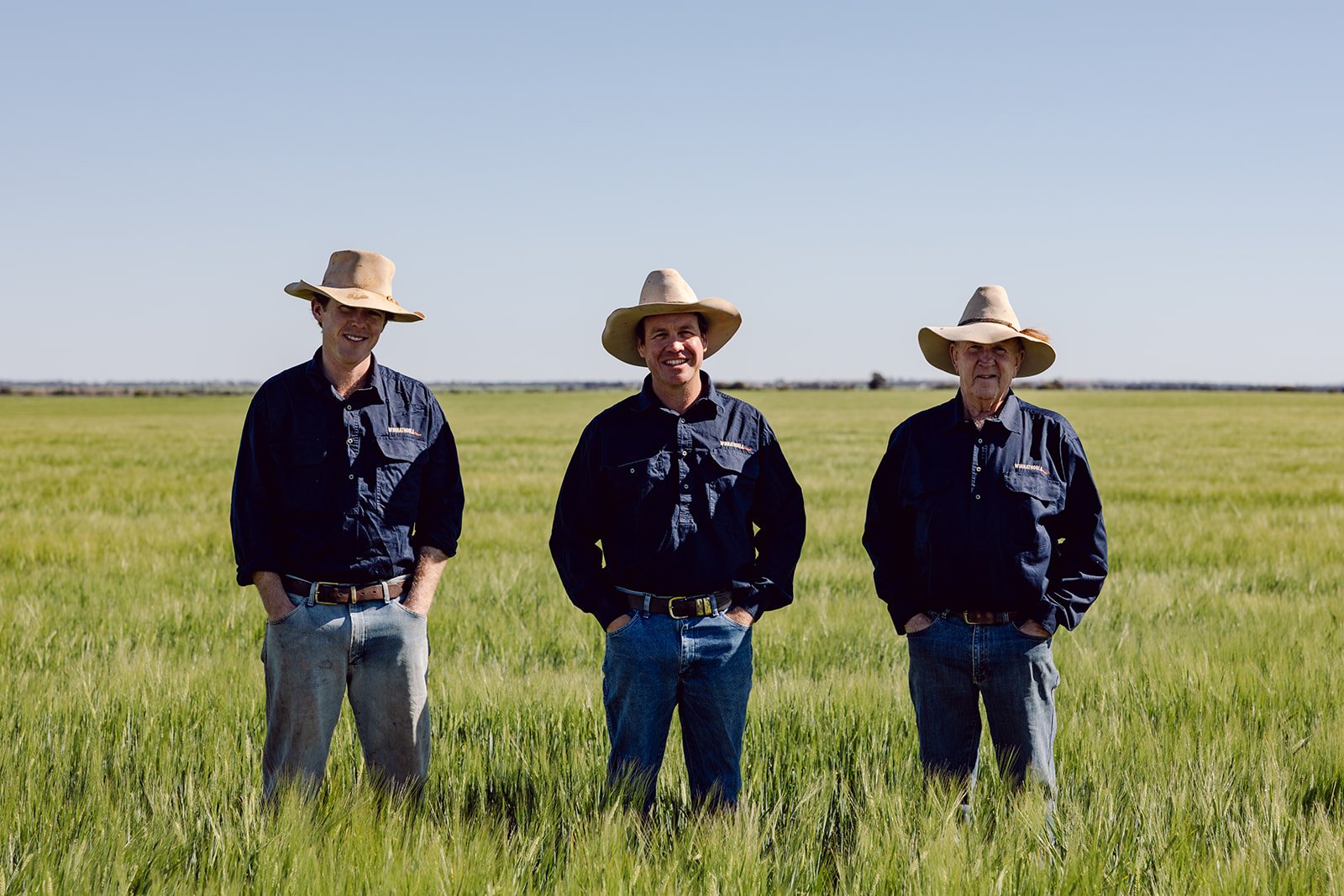 Two men wearing cowboy hats and jeans walk together along a dirt pathway at a cattle farm, with cattle behind metal fences under a clear sky.