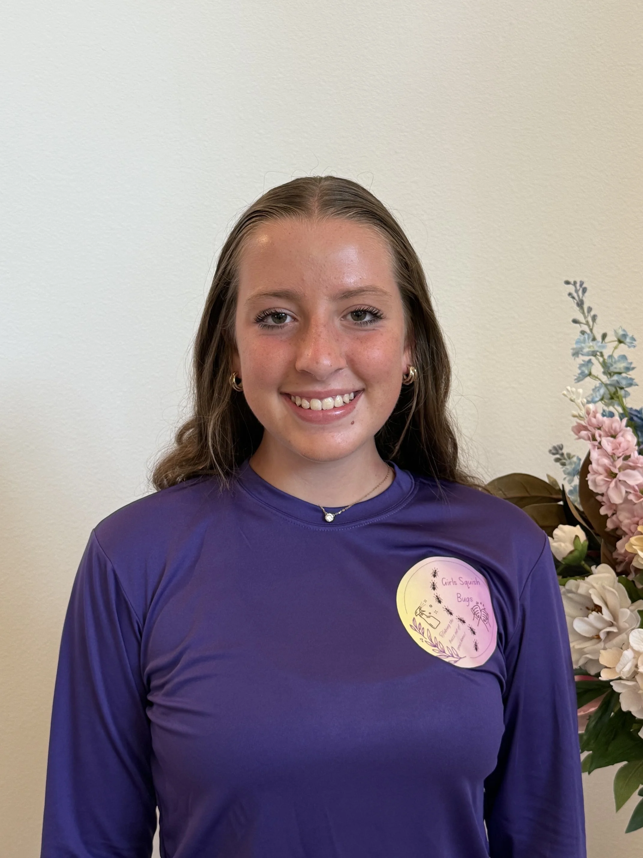 Young woman with curly brown hair, wearing a purple sports shirt, standing indoors with white walls and flowers on her right side, smiling at the camera.