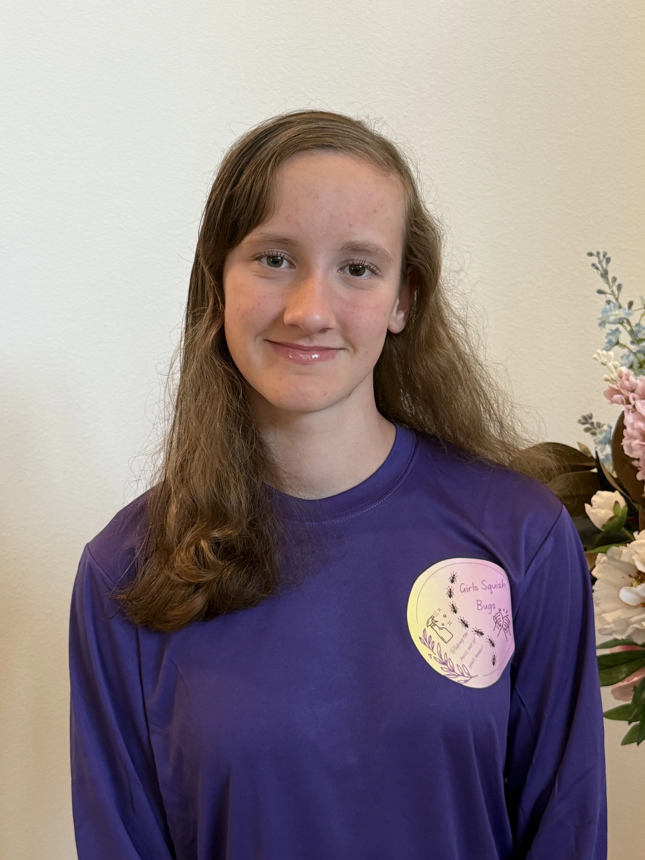 A young girl with long, wavy brown hair and light skin, wearing a purple shirt, standing against a plain light-colored wall with a small grouping of flowers on the right side, and a yellow sticker on her shirt that reads "Girls Squish Bugs."