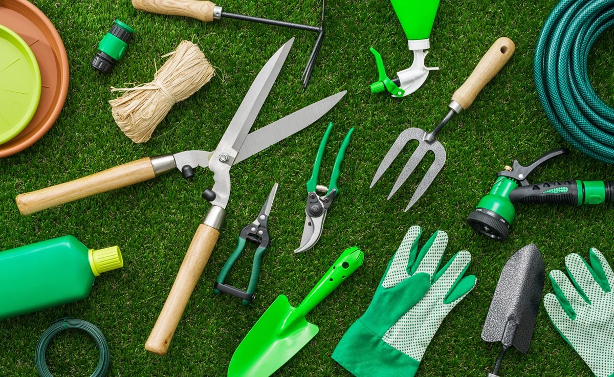 Various gardening tools and supplies on grass, including pruning shears, a trowel, gloves, a garden hose, and a watering nozzle.