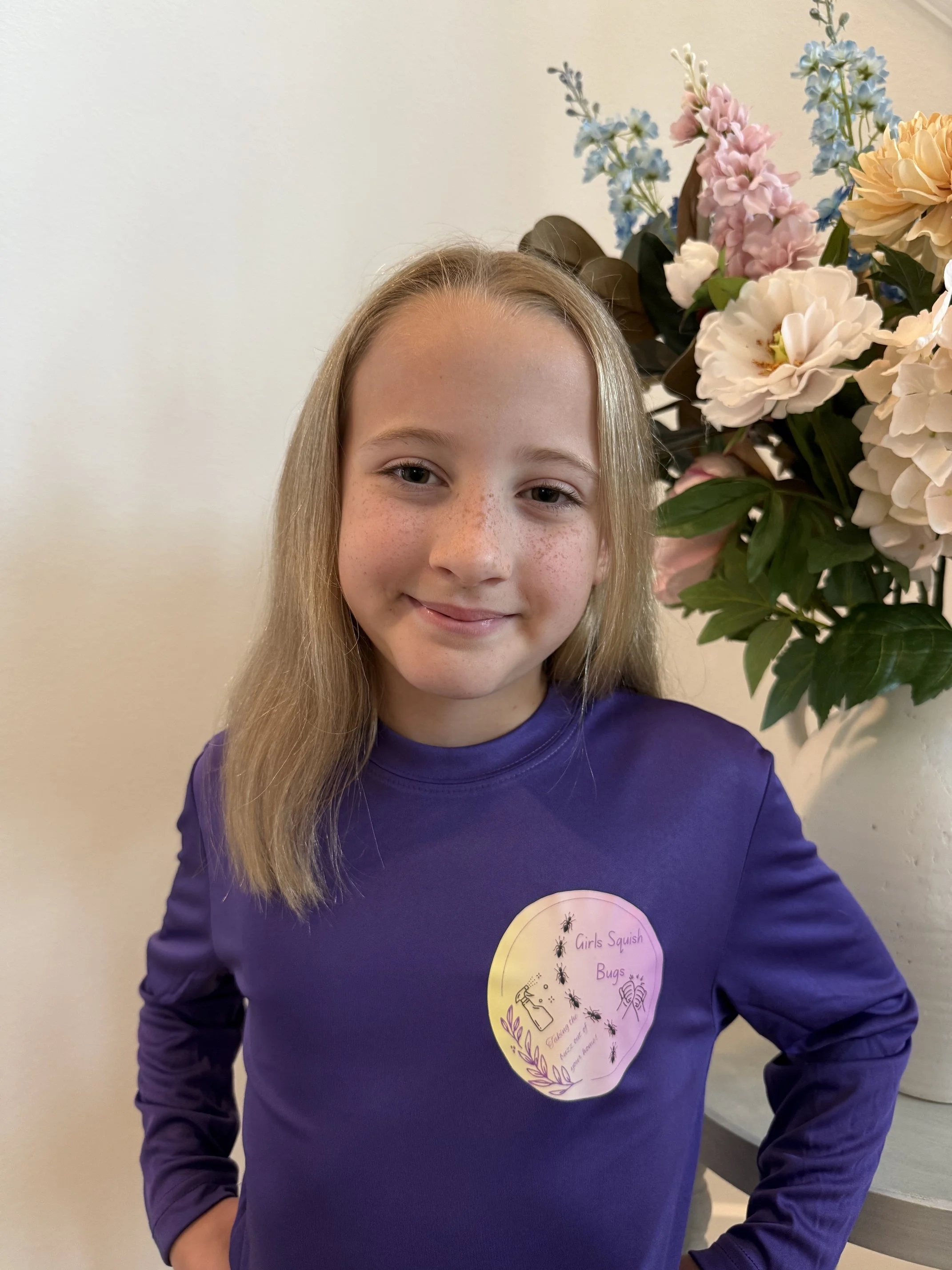 A smiling girl with long blonde hair wearing a purple shirt with a circular badge on it. The badge has text about squish bugs and illustrations of bugs and a spray bottle on it. She is standing in front of a beige wall with a large bouquet of colorful flowers, including white, pink, and blue blooms, to her right.