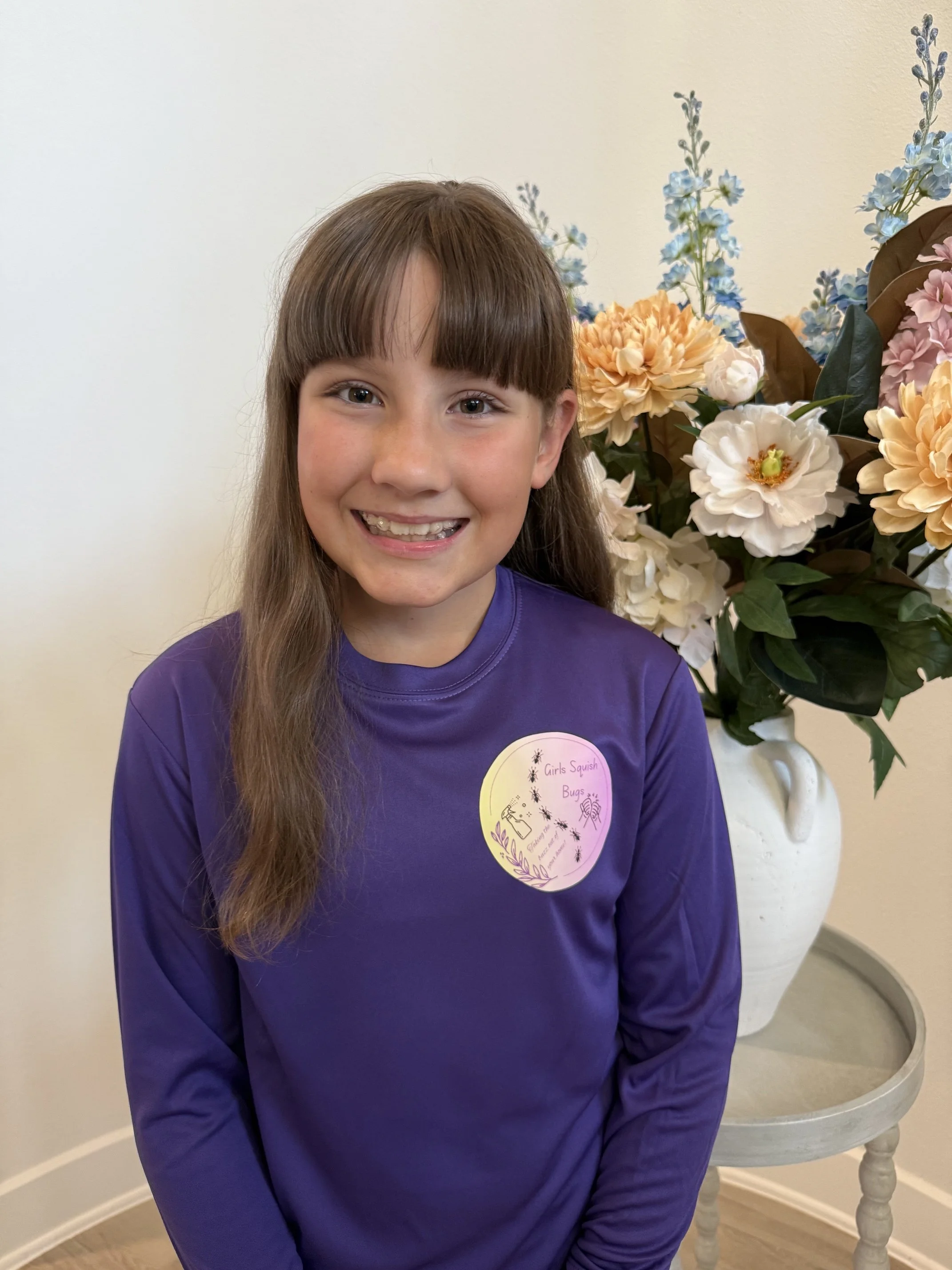 Young girl with long brown hair and bangs, smiling, wearing a purple shirt with a sticker that reads 'Girls Squish Bugs.' She is standing next to a large bouquet of flowers in a white vase, including white, pink, and blue blooms, against a plain light-colored wall.