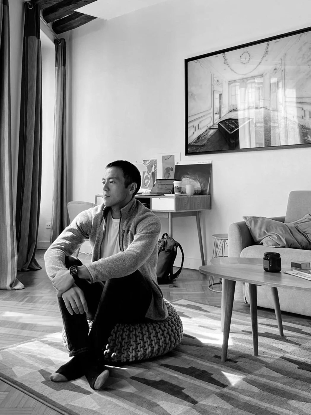A young man sits on a knitted pouf in a living room with large windows and curtains, looking to the side. The room has a sofa, a coffee table with a jar and some items, and a wall with a large framed photograph of an empty room with a piano.