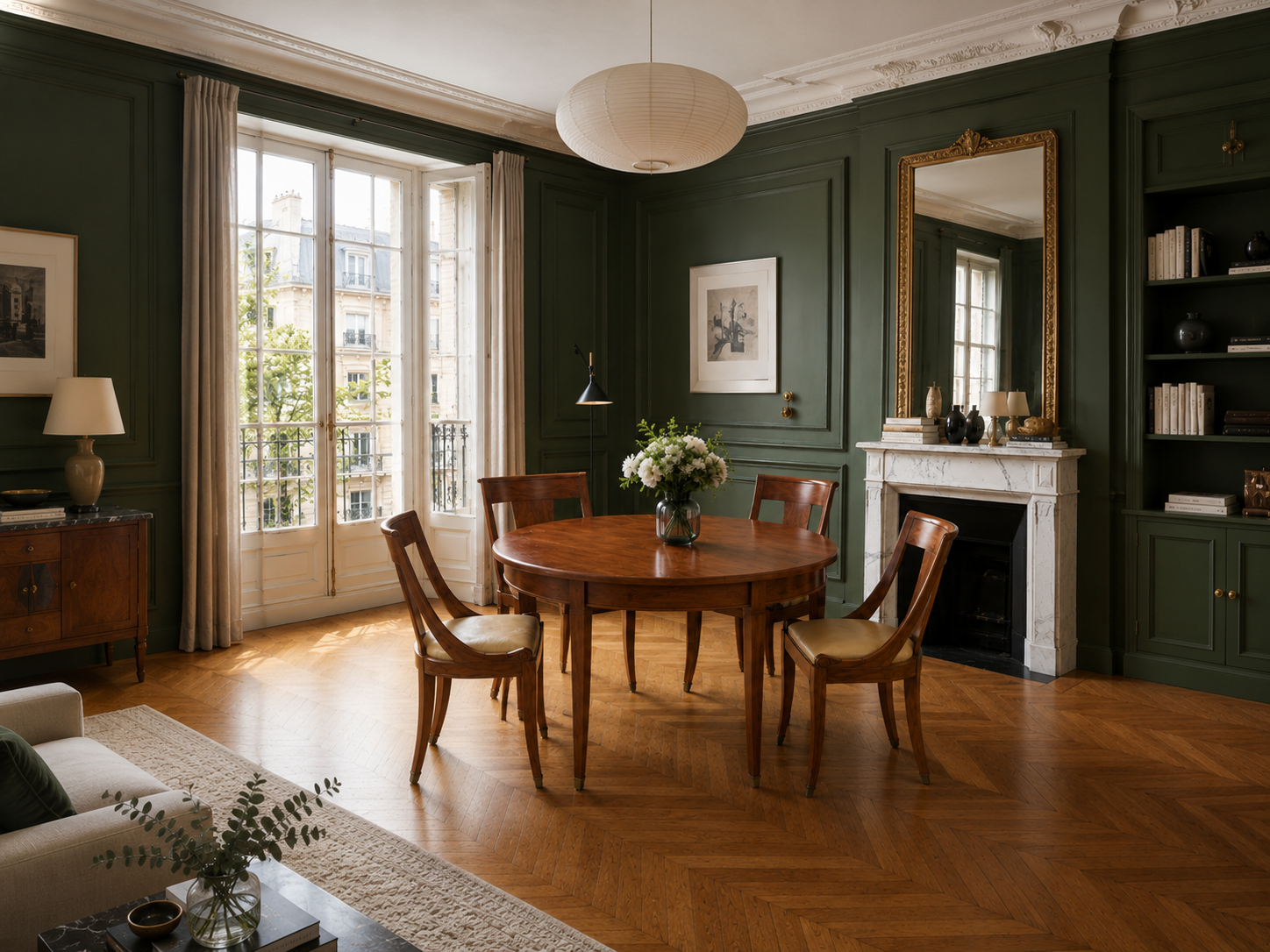 A dining room with dark green paneled walls, a round wooden table with four matching chairs, a vase of white flowers on the table, a marble fireplace with decorative objects, and large windows with beige curtains letting in natural light.
