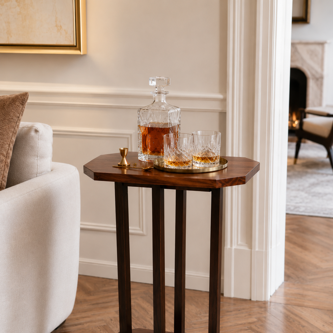 Elegant living room corner with a French Art Deco Style wooden side table holding a crystal decanter with amber liquor, two matching glasses, a gold jigger, and a gold spoon, next to a beige sofa with a decorative pillow.