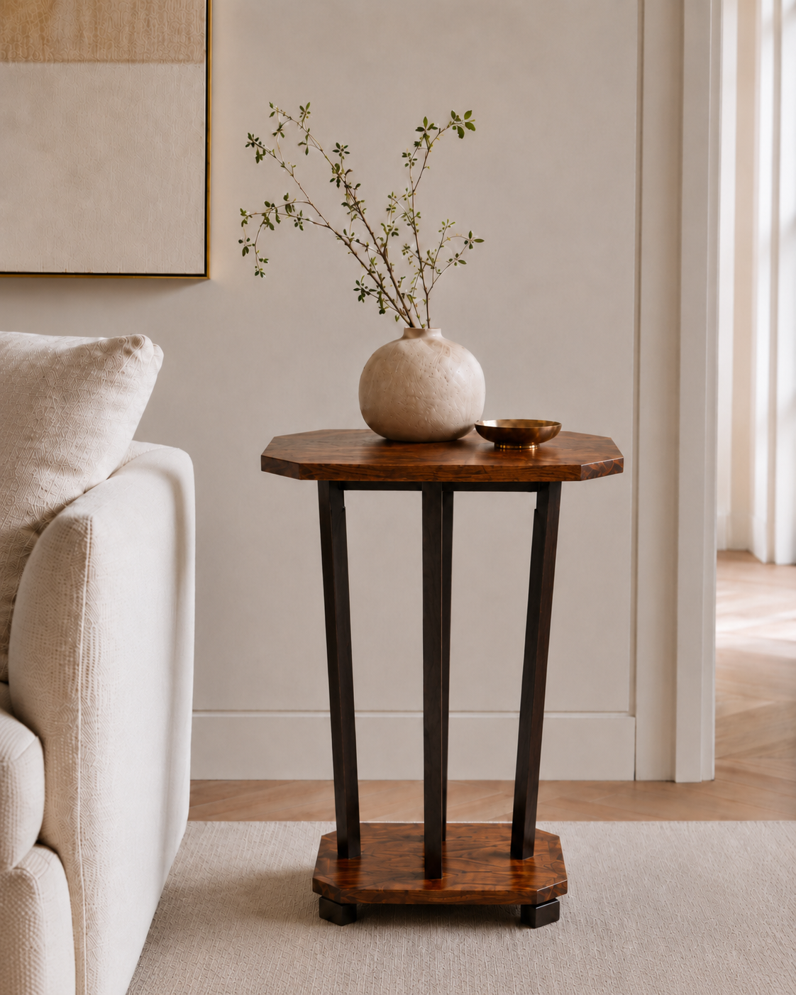 A living room with a small wooden side table holding a white ceramic vase with branches and a small decorative bowl. Part of a white upholstered sofa is visible on the left, and the background features a wall with artwork and a doorway to another room.