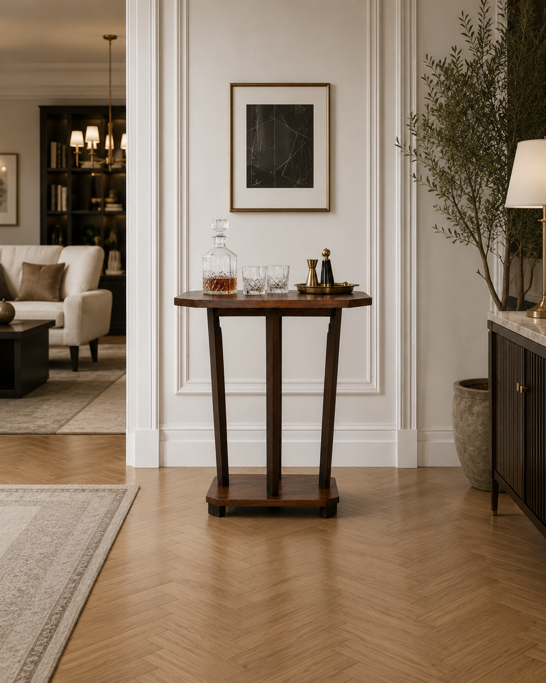 French Art Deco Style wooden console table with a glass decanter, two glasses, and decorative bottles, against a white paneled wall with a framed black artwork. Part of a living room with a beige sofa, bookshelf, and chandelier visible.