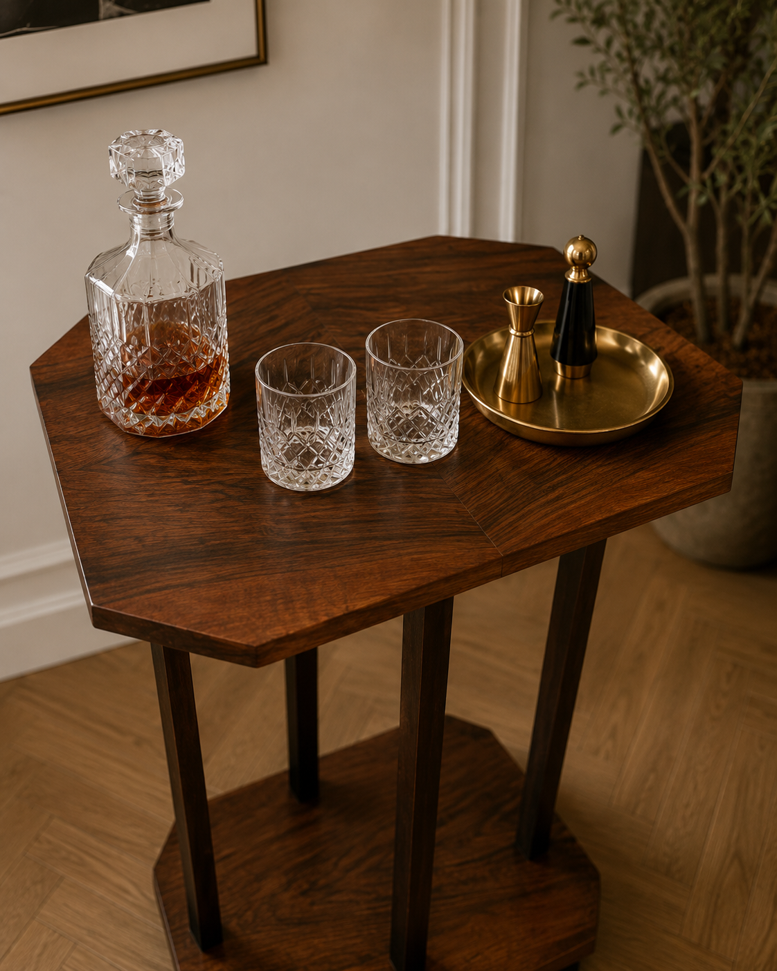 A wooden bar cart holding a crystal decanter of whiskey, two cut glass whiskey glasses, and a gold and black cocktail shaker with a gold tray, set against a white wall with a framed picture and a potted plant in the background.