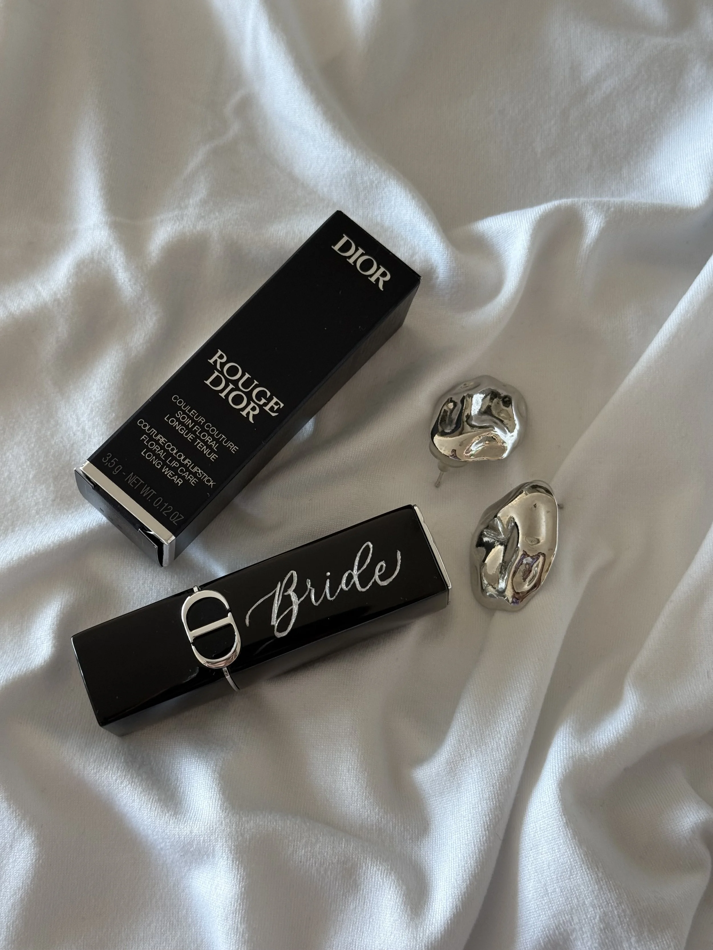A black Dior lipstick box, a black item with the word 'Bride' in cursive, and metallic decorative objects on a white satin fabric.
