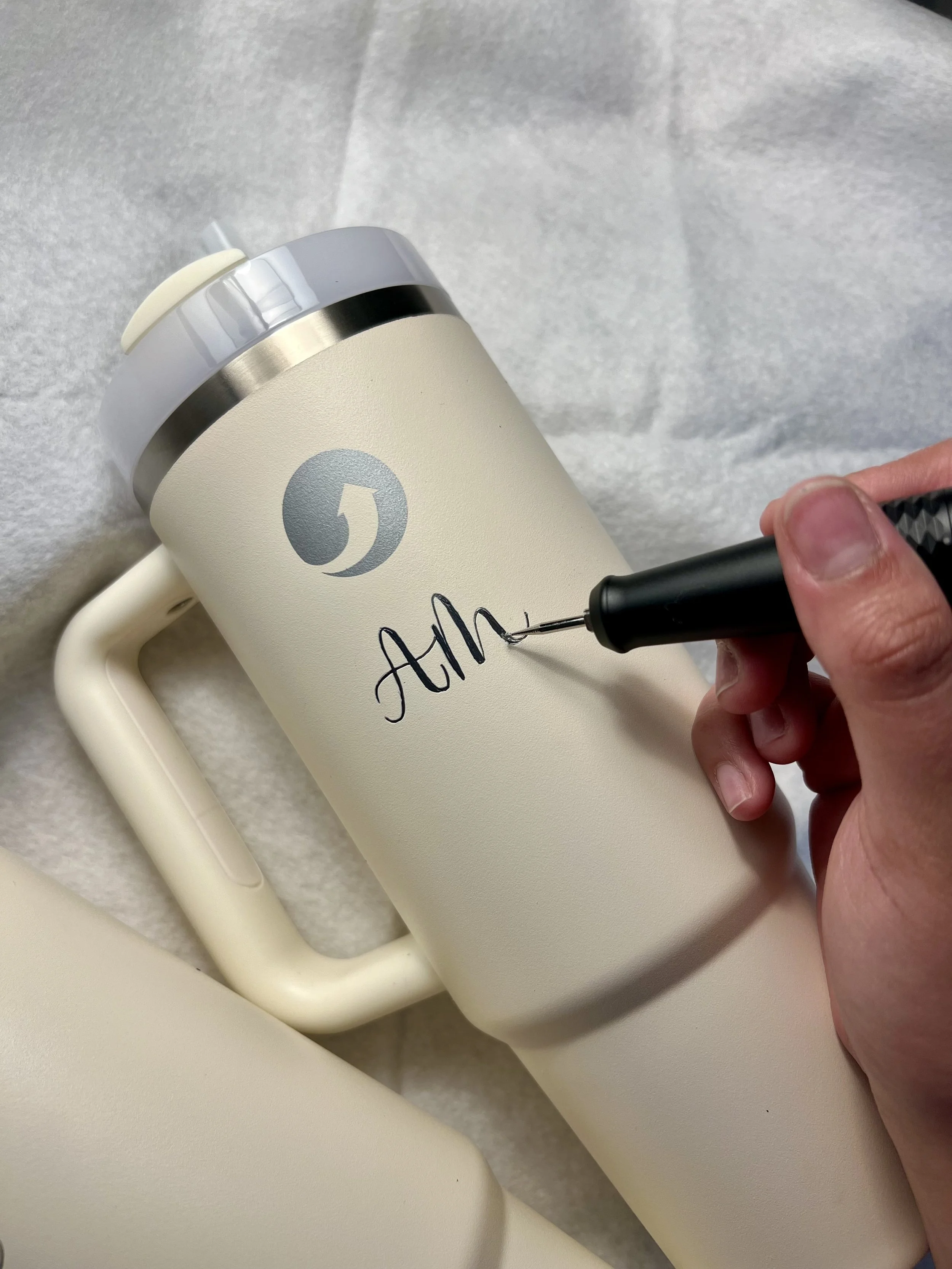 A person is signing their name on a beige travel mug with a black marker. The mug has a silver band near the top and a cream-colored lid. The mug is placed on a white surface, and part of a beige item or paper is visible in the background.