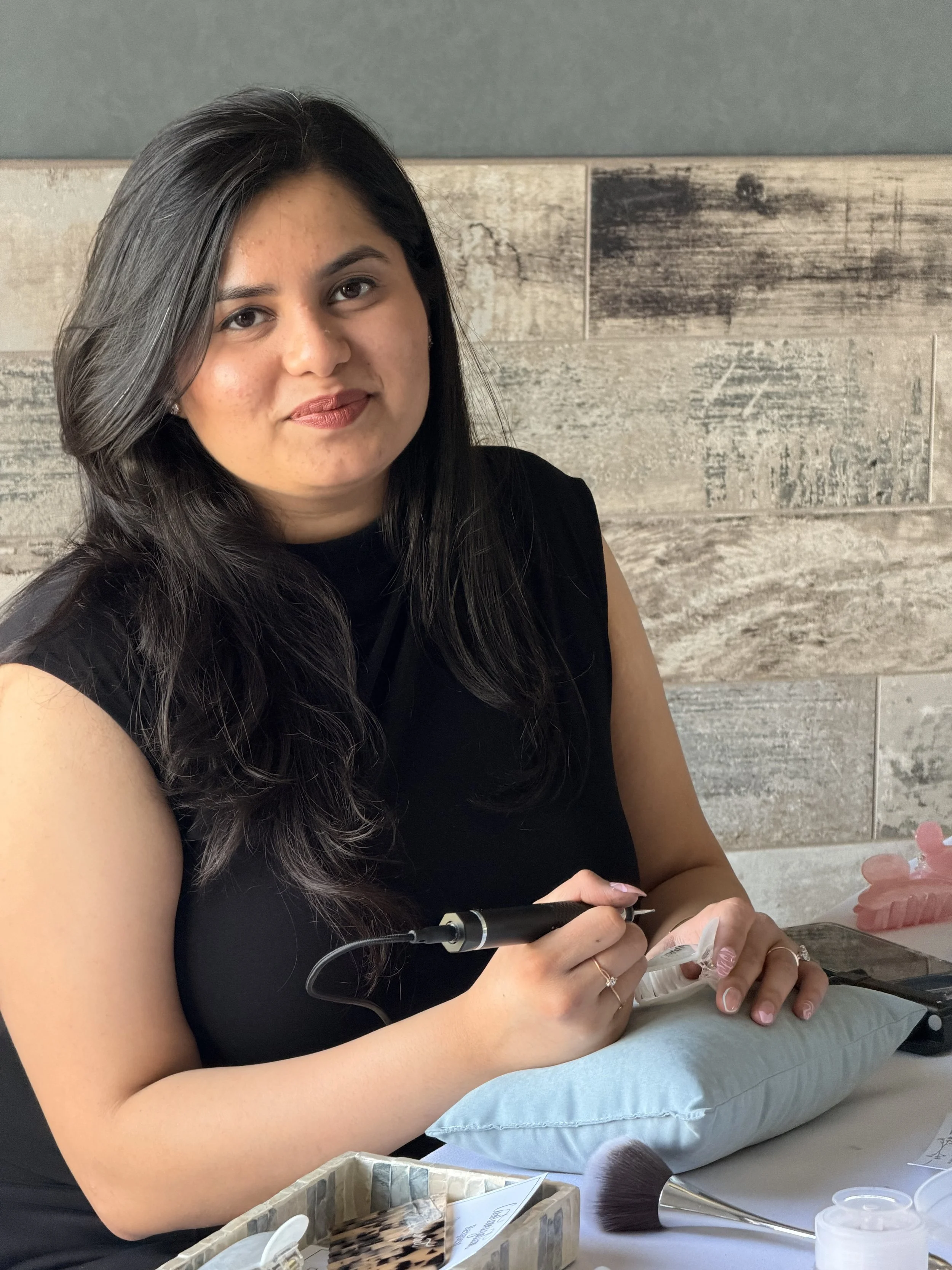 A woman with long dark hair, wearing a black shirt, seated at a table with makeup tools and products, holding a makeup brush.