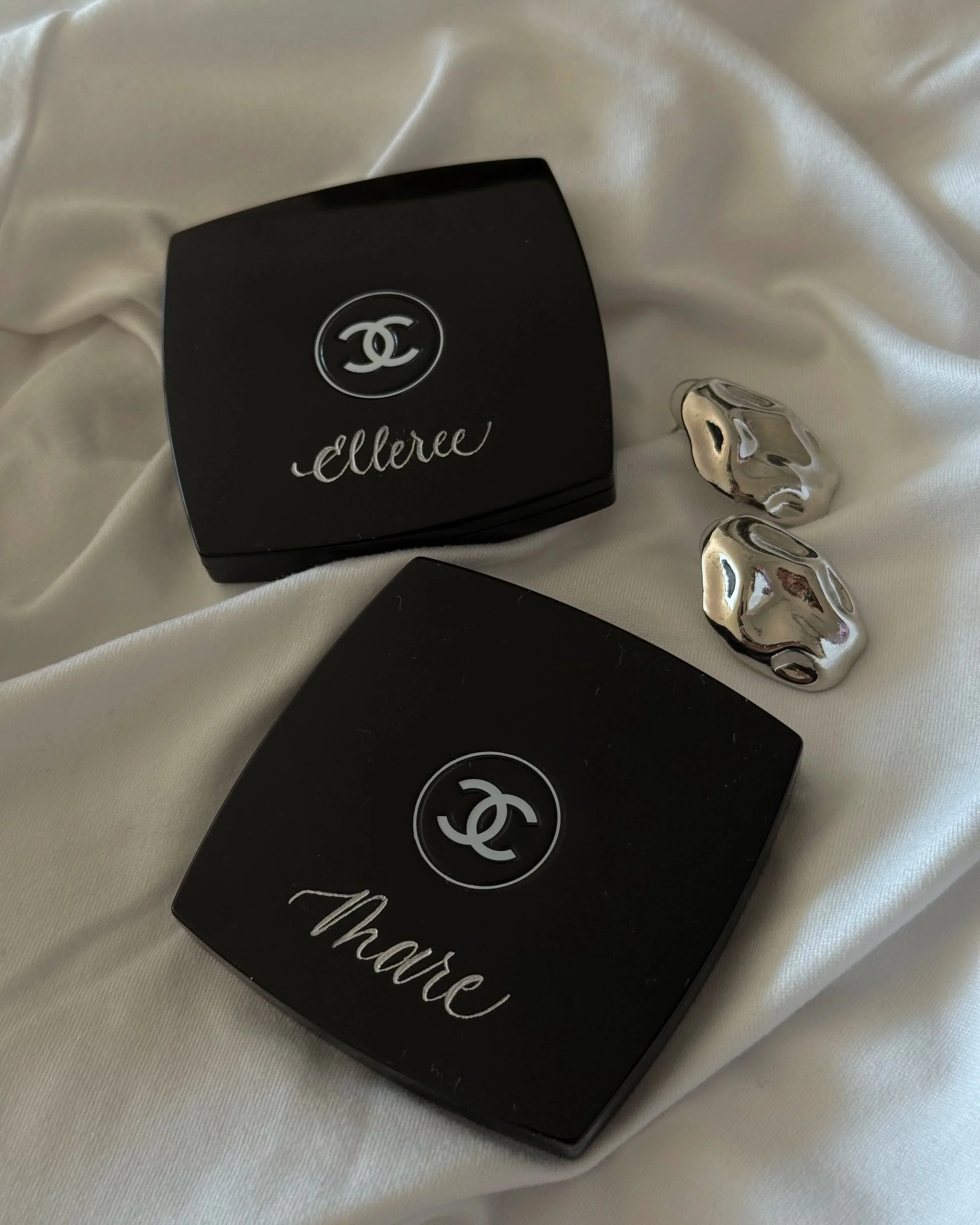 Black compact mirror with the Chanel logo on the lid, placed on white satin fabric along with three metallic silver abstract-shaped objects