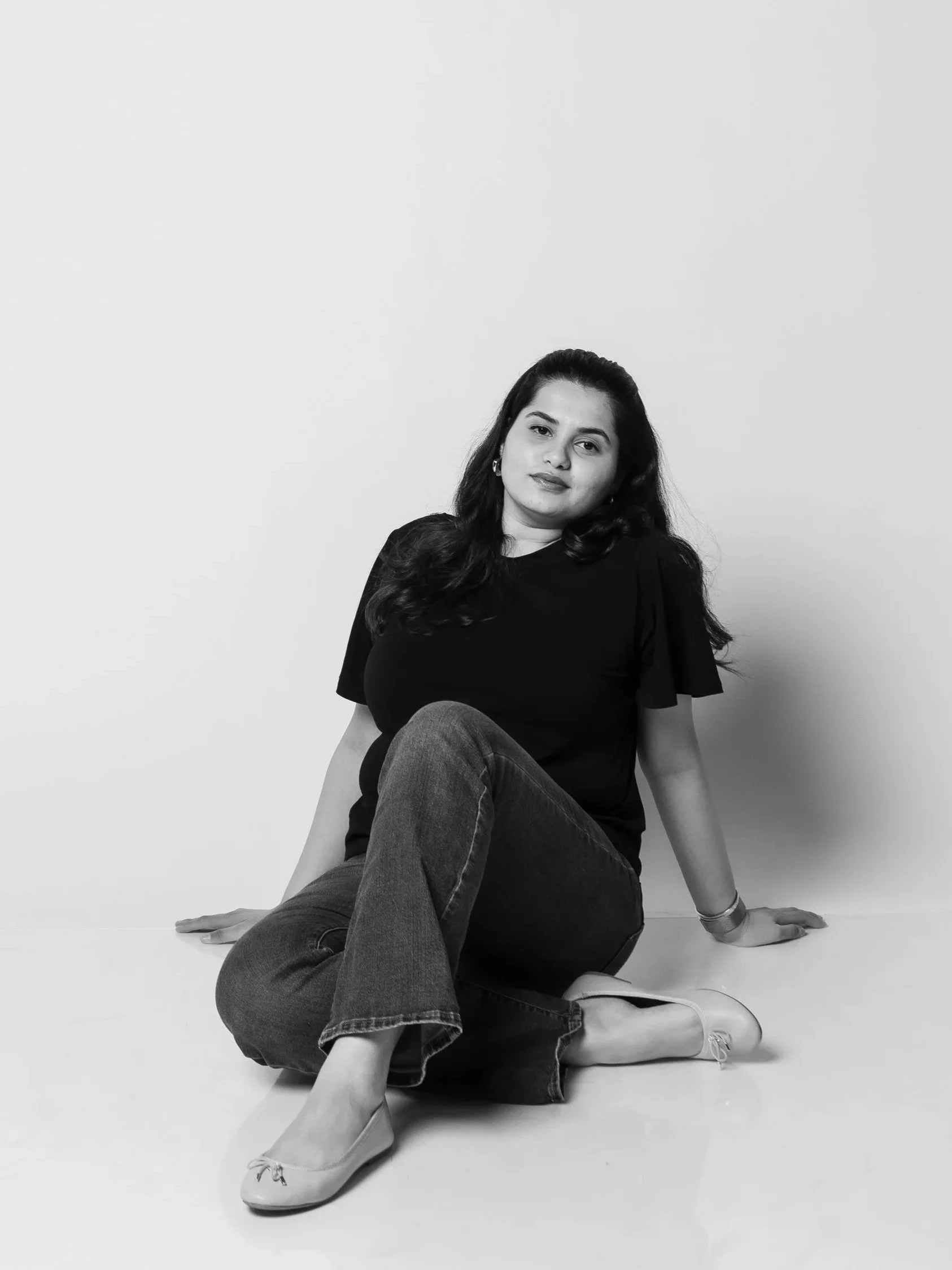 A young woman with long dark hair sitting on the floor against a plain wall, wearing a black top, jeans, and light-colored shoes, looking directly at the camera.