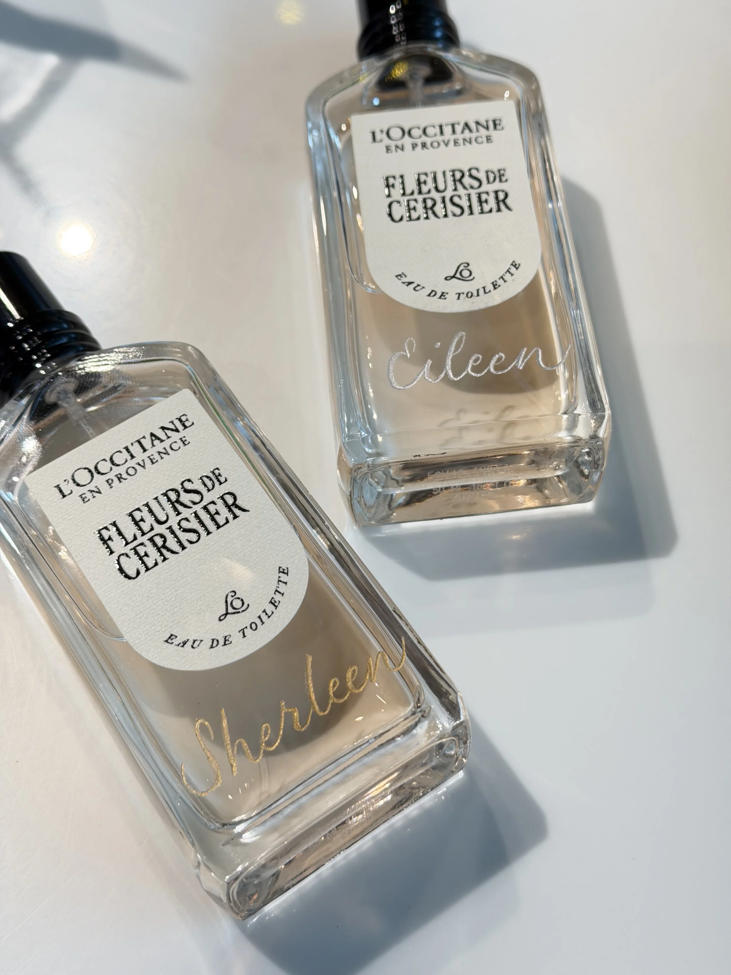 A clear rectangular perfume bottle labeled 'Fleurs de Cerisier' (Cherry Blossom) with a black cap, placed on a white surface, with another similar bottle partially visible.