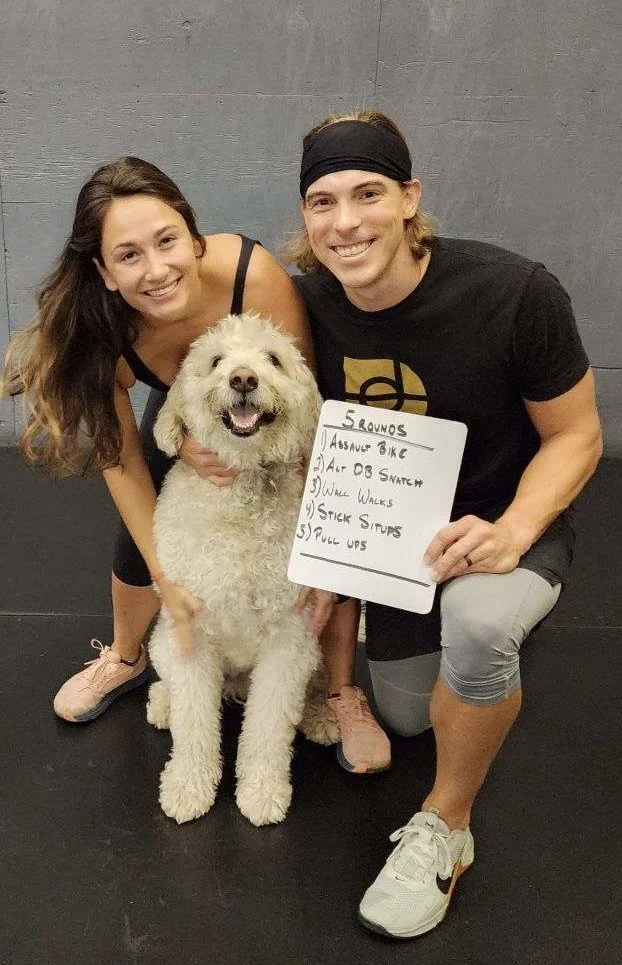 Two people smiling and kneeling beside a large fluffy dog indoors. One person is holding a whiteboard with a workout list including exercises like Assault Bike, DB Snatch, and Pull Ups.