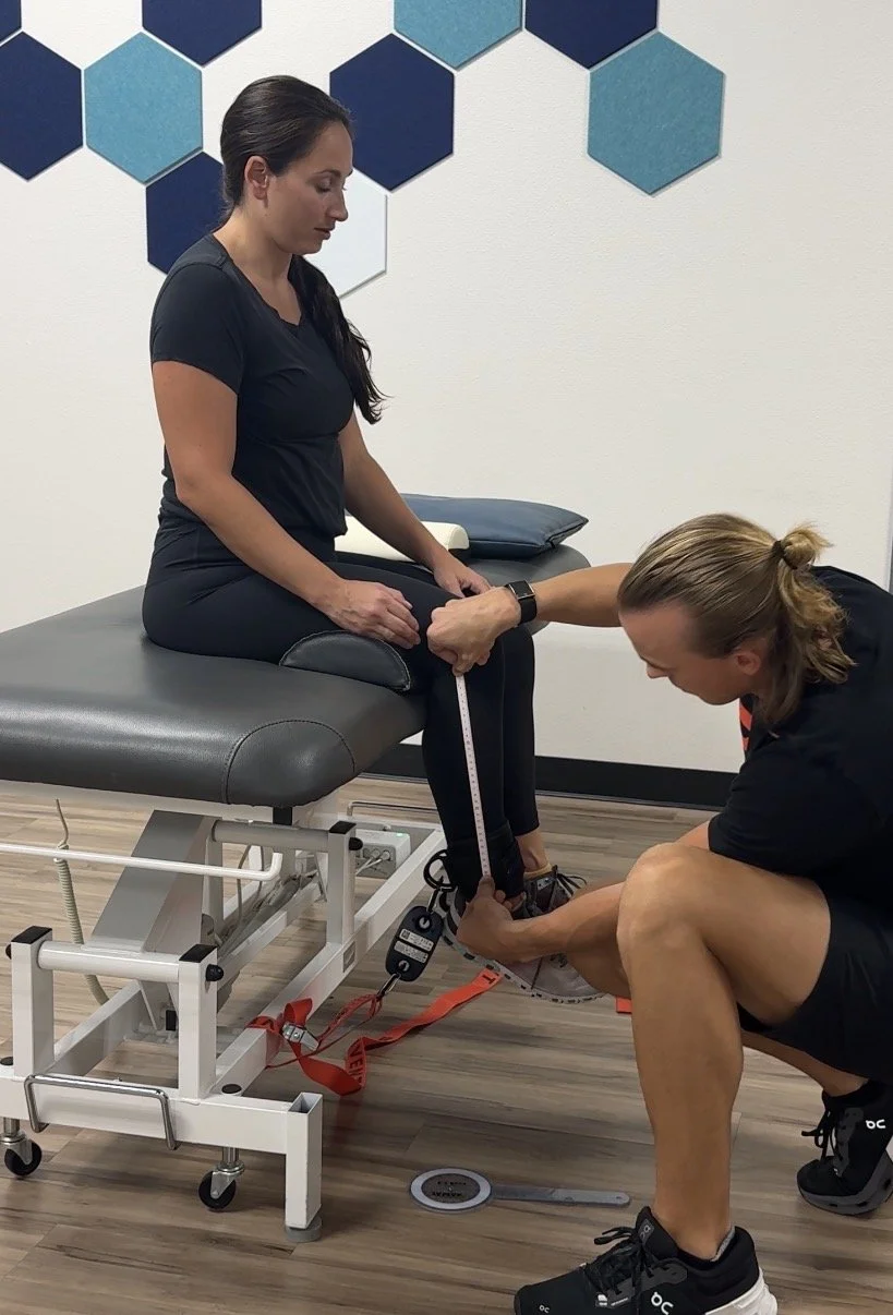A woman sitting on a treatment table with her leg extended, while a physical therapist measures her lower leg with a tape measure, in a clinical setting with a geometric blue hexagon pattern on the wall.