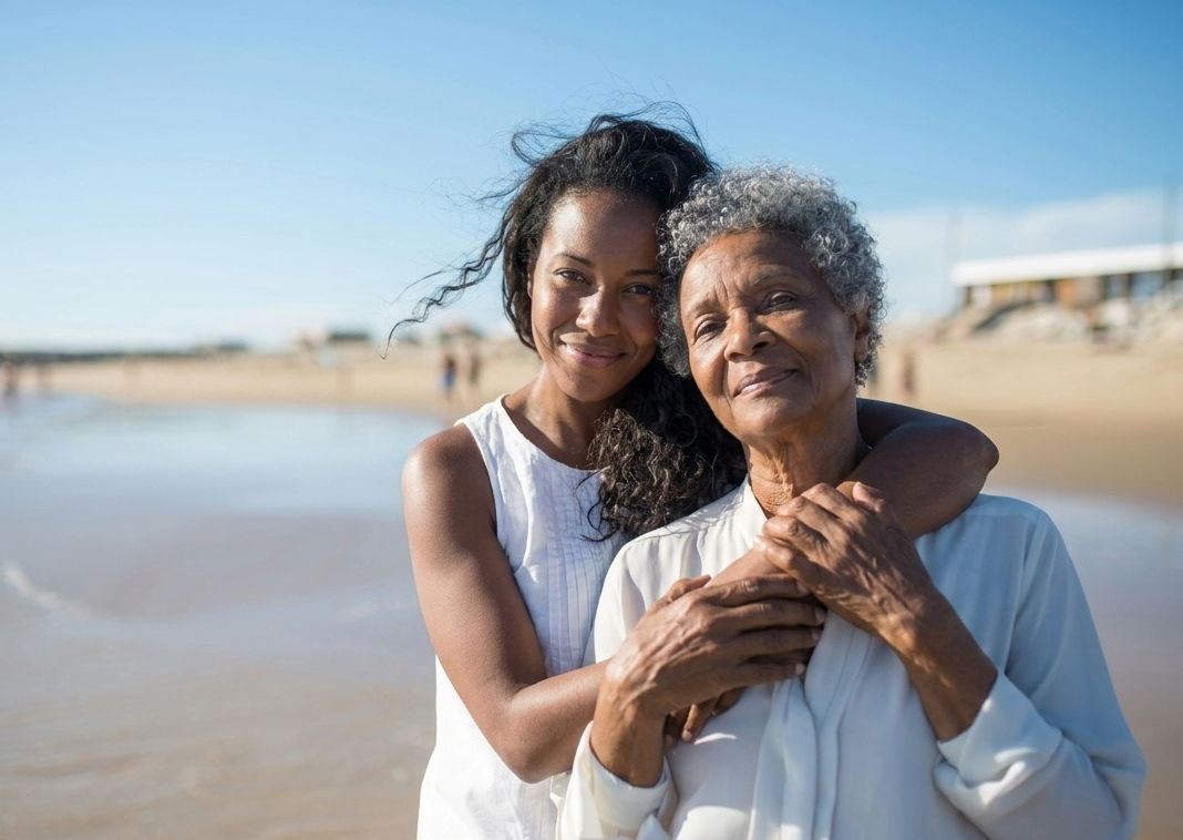 A young woman and an elderly woman hugging on a beach with blue sky and ocean in the background.