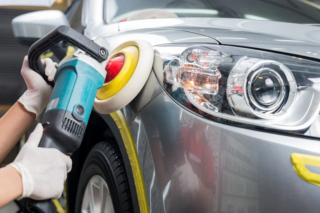 A person polishing a gray car's front fender with a power buffer, wearing white gloves.