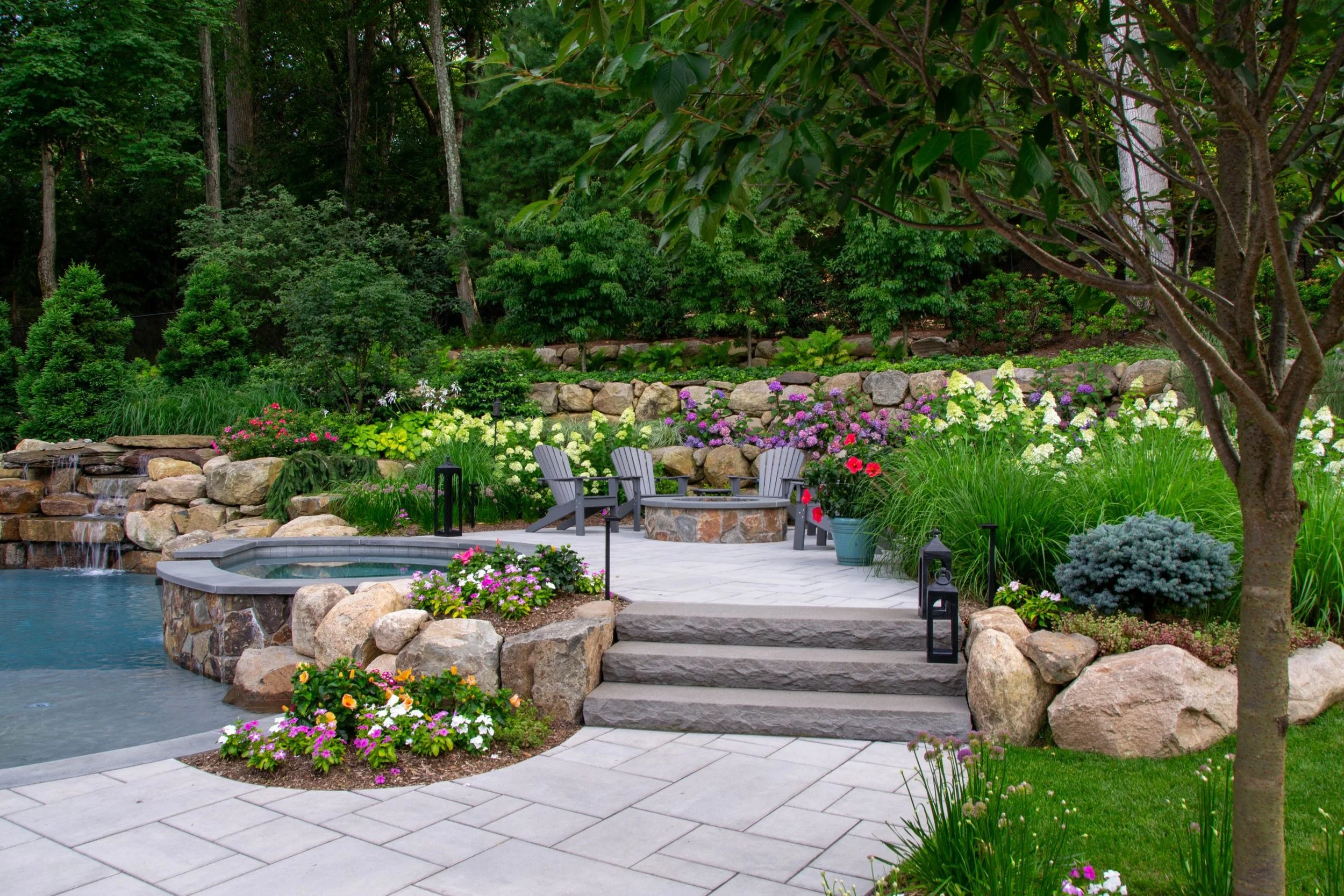 Backyard garden with stone steps, potted plants, flower beds, a fire pit with Adirondack chairs, water features, and lush greenery surrounded by trees.