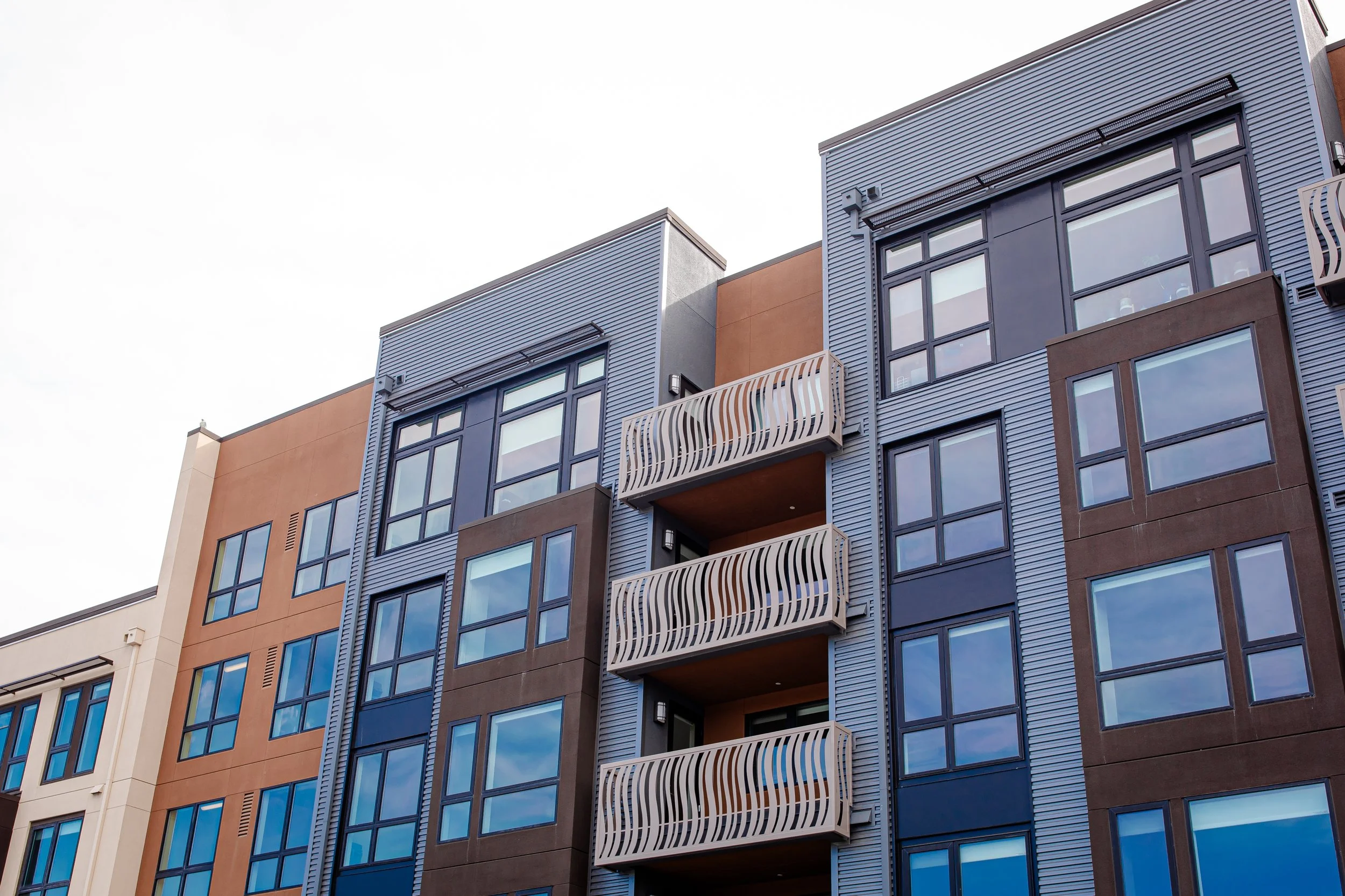 Modern multi-story apartment building with blue, brown, and beige exteriors featuring large windows and unique wavy balcony railings.