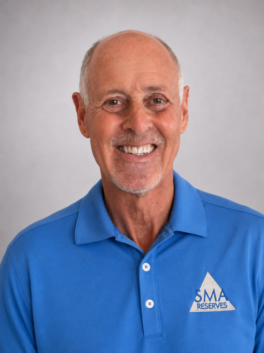 A smiling older man with a bald head and gray facial hair, wearing a blue polo shirt with a logo that reads 'SMA RESERVES'.