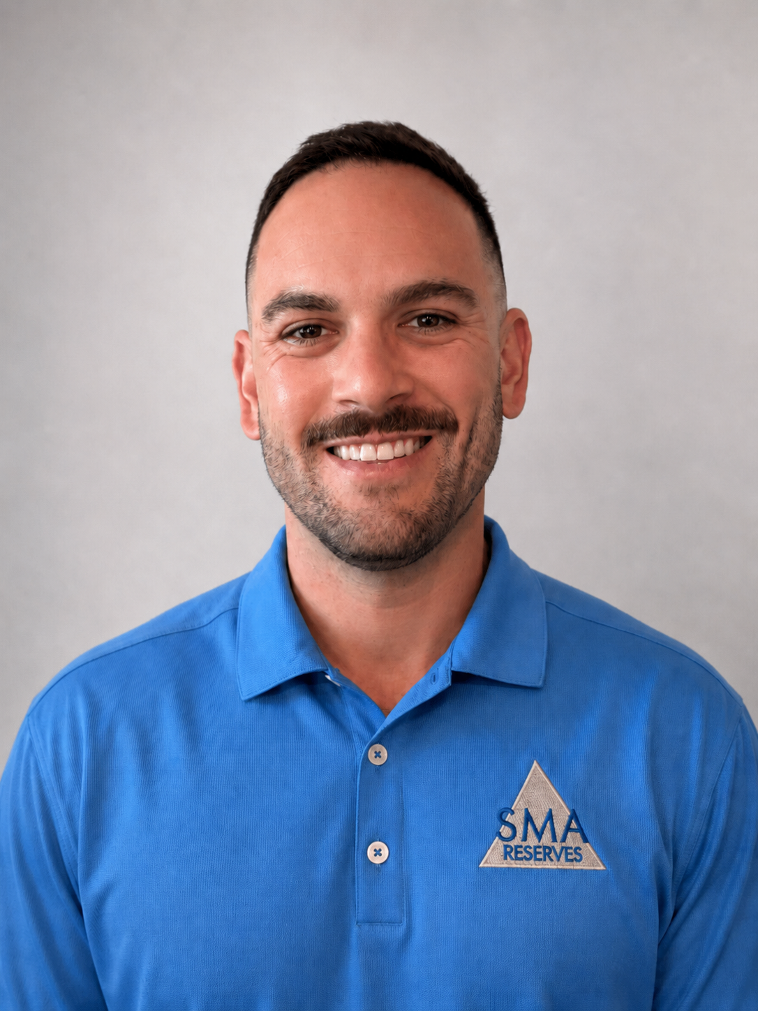 A man with short dark hair and a beard smiling, wearing a blue polo shirt with a logo that reads 'SMA RESERVES' on the chest, standing against a plain gray background.