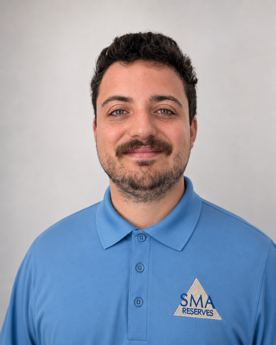 A man with dark brown curly hair, a beard, and mustache, wearing a blue polo shirt with a logo "SMA RESERVES" on the chest, smiling and standing against a plain light gray background.