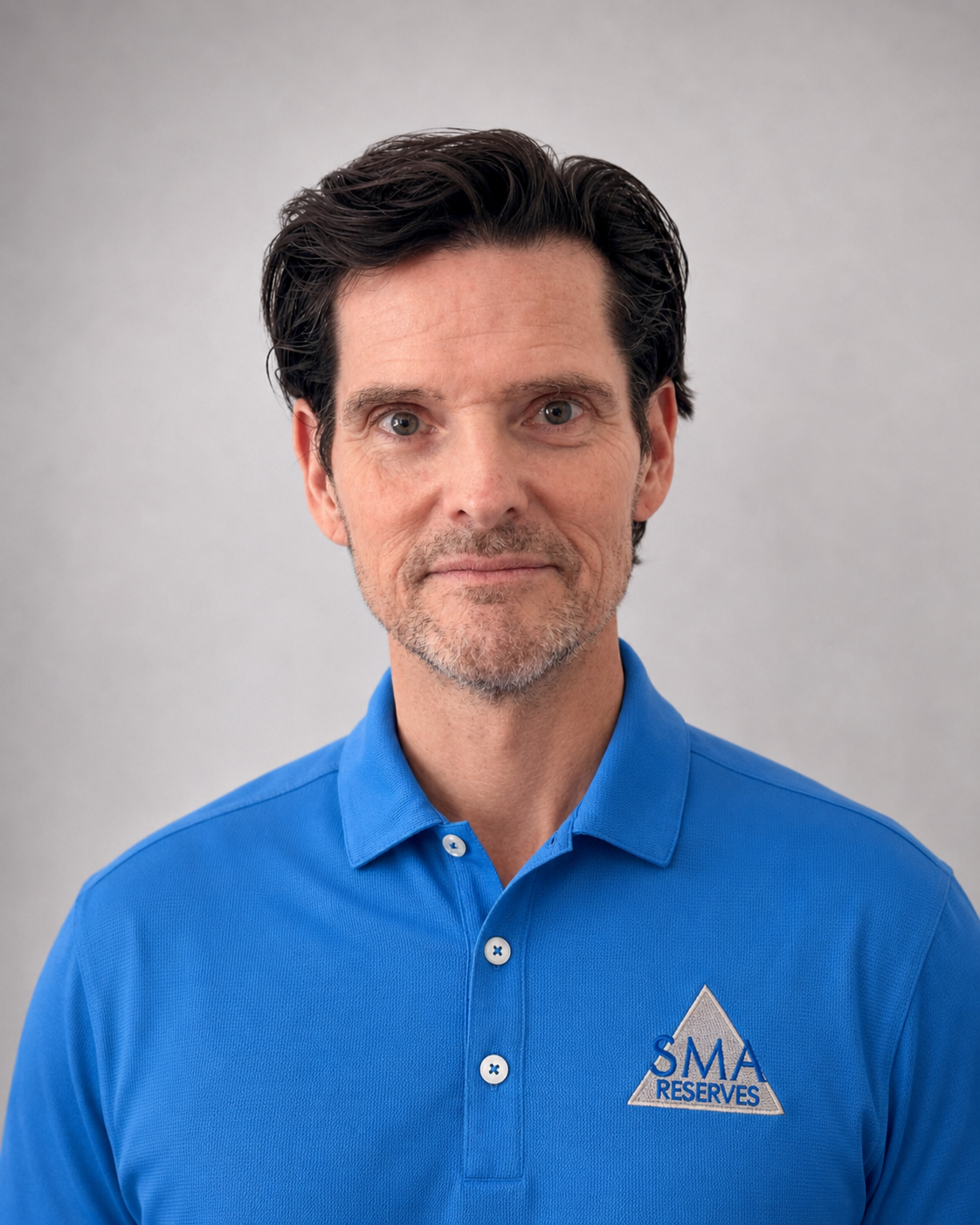 A middle-aged man with dark hair and a beard, wearing a blue polo shirt with a logo that reads 'SMA Reserves,' standing against a plain light gray background.
