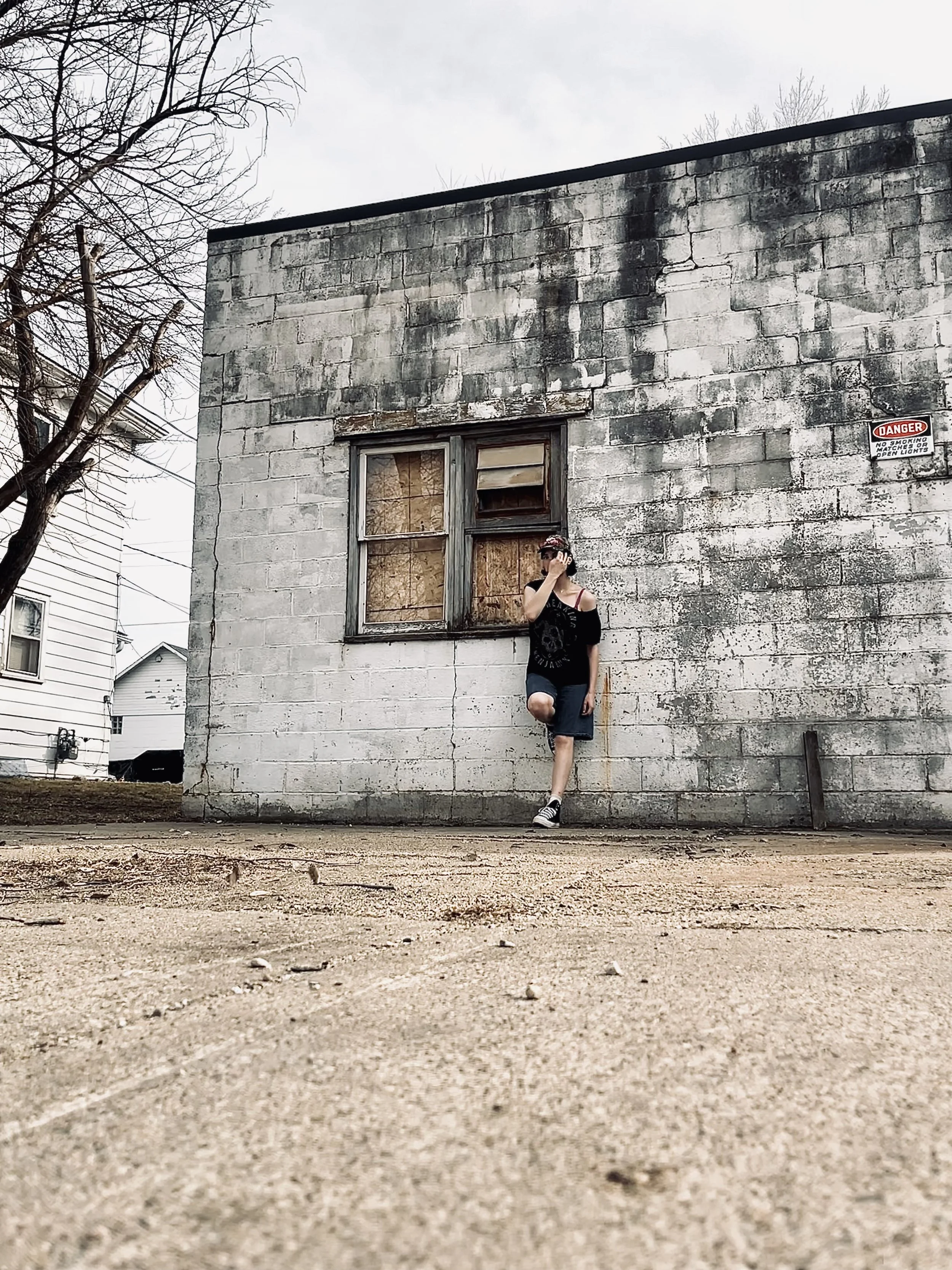 A person standing in front of an old, weathered brick building with boarded-up windows. The person is wearing a black shirt, denim shorts, and sneakers, with one knee bent and resting against the building. Bare tree branches are visible to the left, and a warning sign is attached to the building.