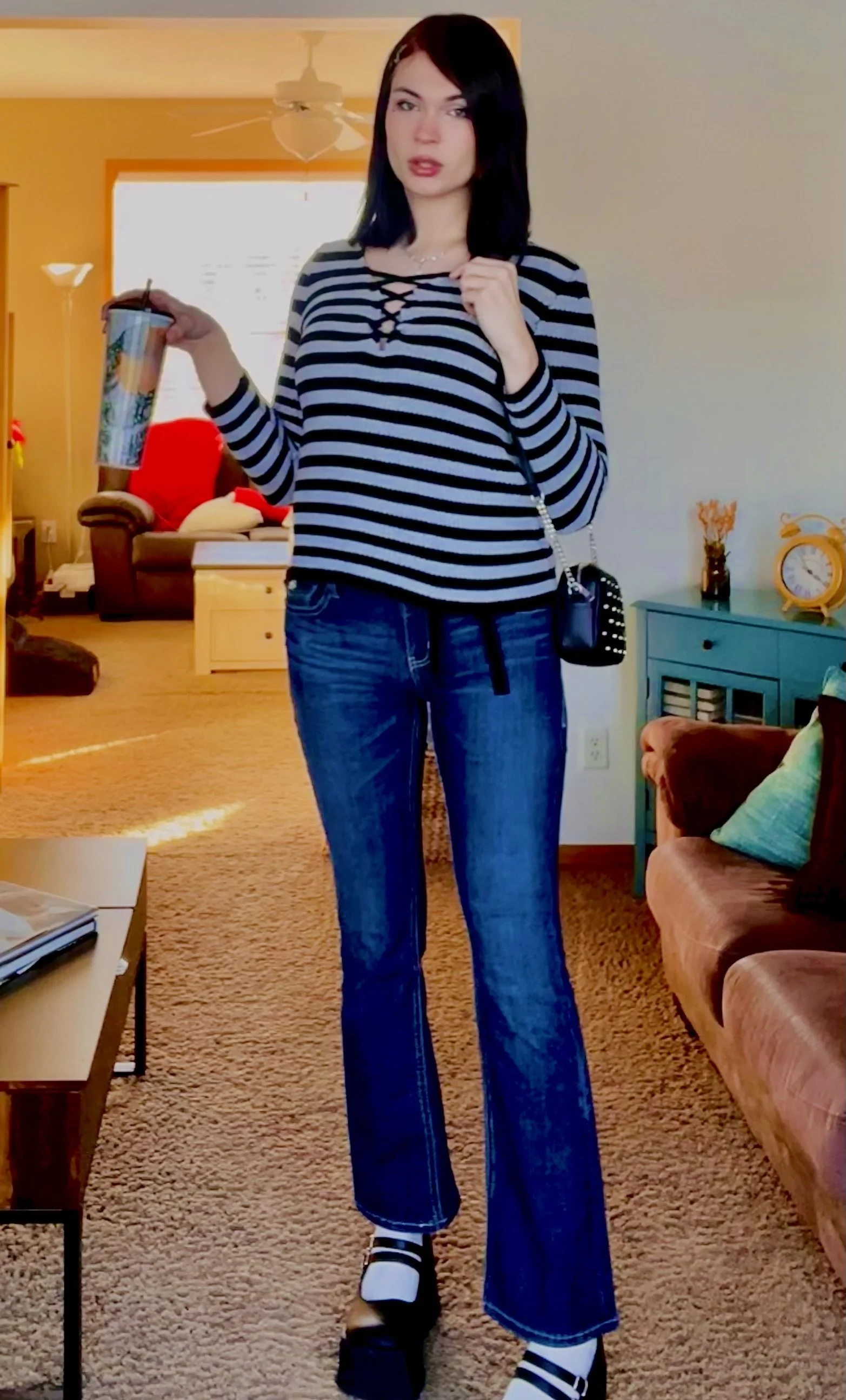 A woman with shoulder-length black hair, wearing a black and white striped long-sleeve top with crisscross detailing at the neckline, blue wide-leg jeans, and black and white striped socks with black shoes, standing in a living room holding a large cup with a straw in her right hand.