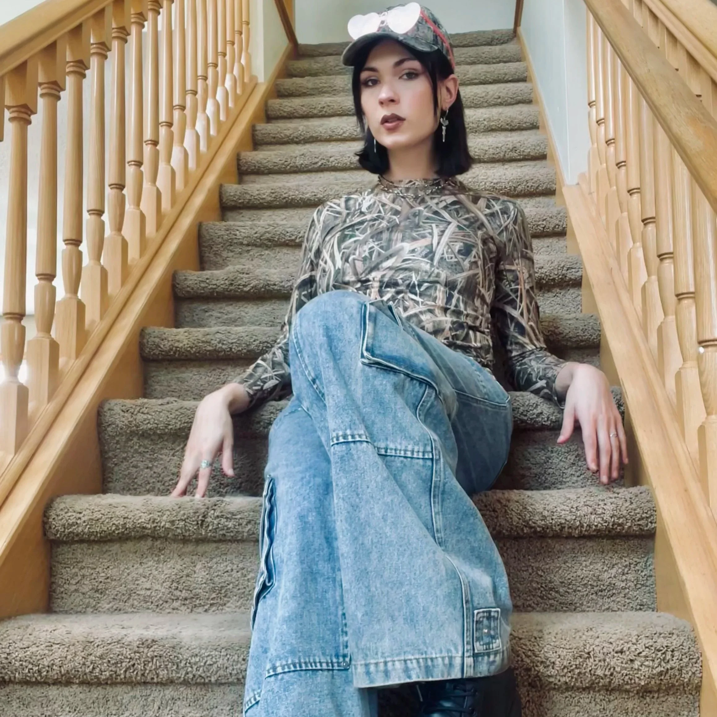 A young woman with black hair sitting on carpeted stairs inside a home, wearing a patterned long-sleeve shirt, jeans, a baseball cap, and sunglasses on her head, with a wooden railing on each side of the staircase.