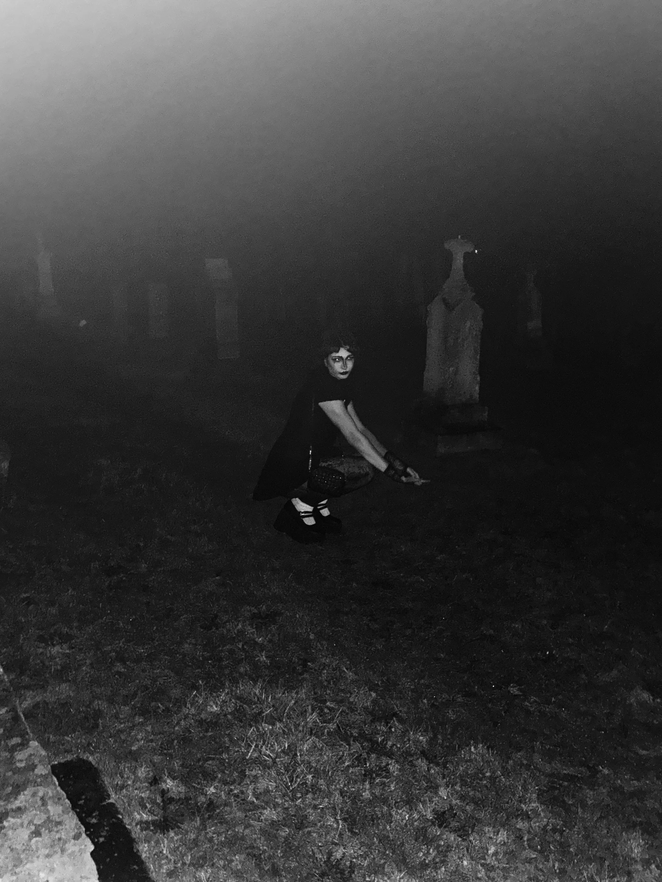 A person with dark hair and dark lipstick squatting on the ground at night in a graveyard, holding a leash, with stone tombstones visible in the background.